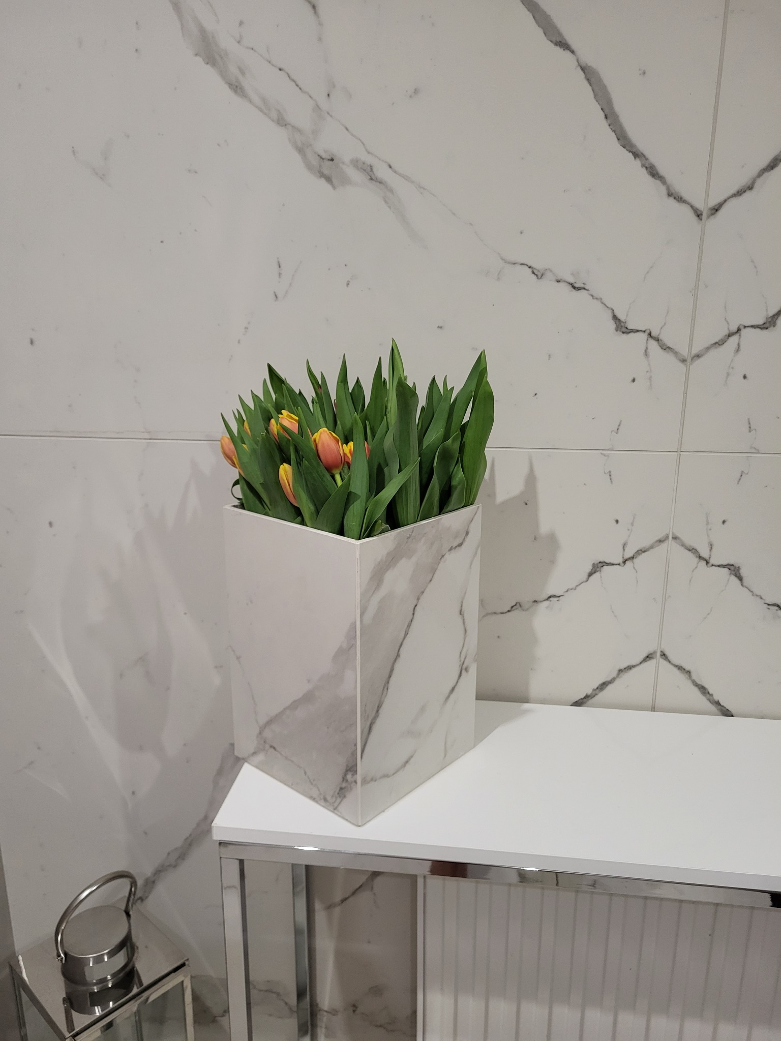 Kwadratowa donica z wzorem marmuru, wypełniona tulipanami, stoi na białym stoliku na tle ściany wyłożonej płytkami imitującymi marmur.