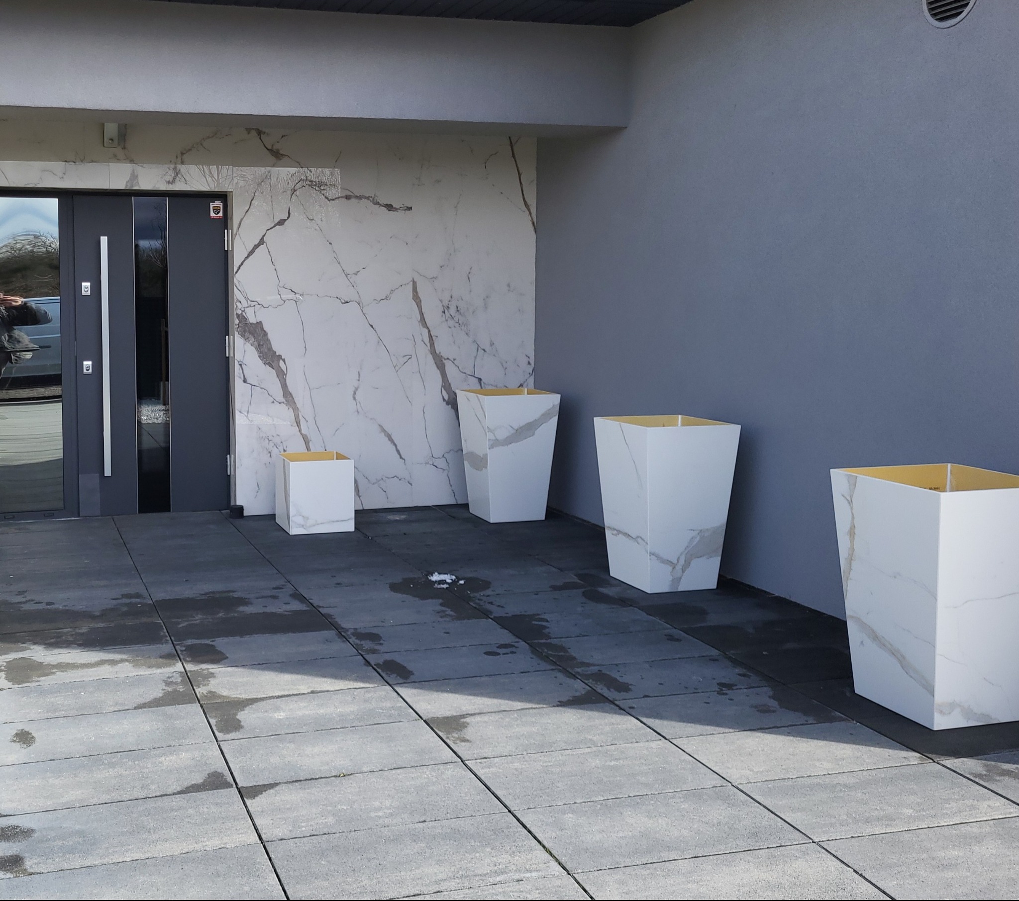 Nowoczesne wejście do domu z szarymi płytami tarasowymi, białymi donicami z motywem marmuru i elewacją z płyt imitujących marmur przy drzwiach.