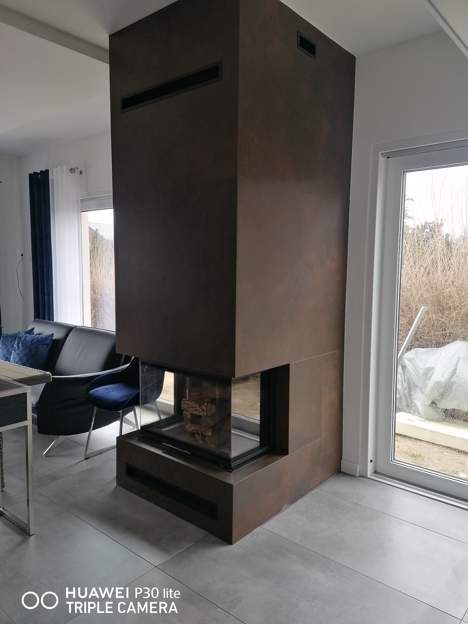 Nowoczesny, obudowany kominek z ciemnobrązowych płyt w salonie, z widocznym paleniskiem i ułożonymi w nim kawałkami drewna, z prostokątnymi otworami wentylacyjnymi w górnej części obudowy, na tle...