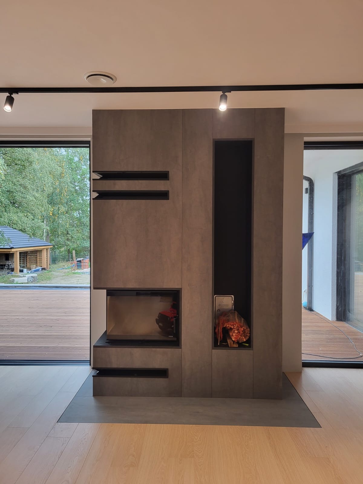 Nowoczesny szary kominek z wkładem i wnęką na dekoracje, oświetlony punktowymi lampami sufitowymi, widok na taras i ogród przez przesuwne okno.