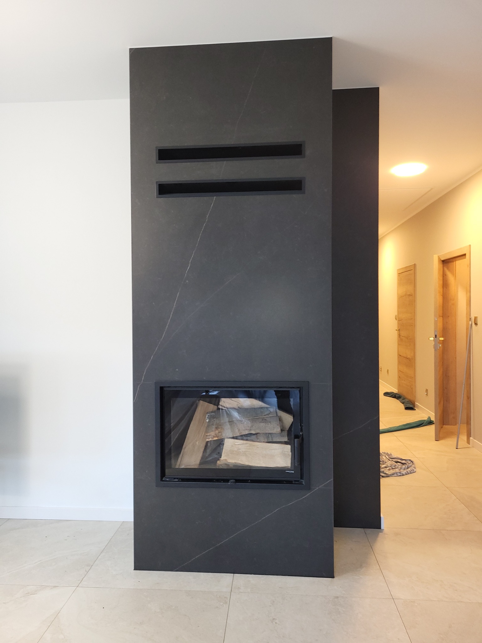 Nowoczesny kominek z obudową z ciemnego kamienia z wyraźnym użyleniem, z dwoma poziomymi wnękami nad paleniskiem, z drewnem w środku, na tle białej ściany i jasnej podłogi z płytek.