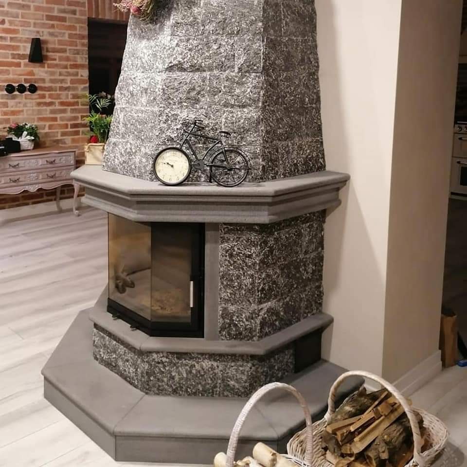 Szary, narożny kominek z kamienną obudową, ozdobiony zegarem w kształcie roweru, z koszem drewna obok, w jasnym wnętrzu.