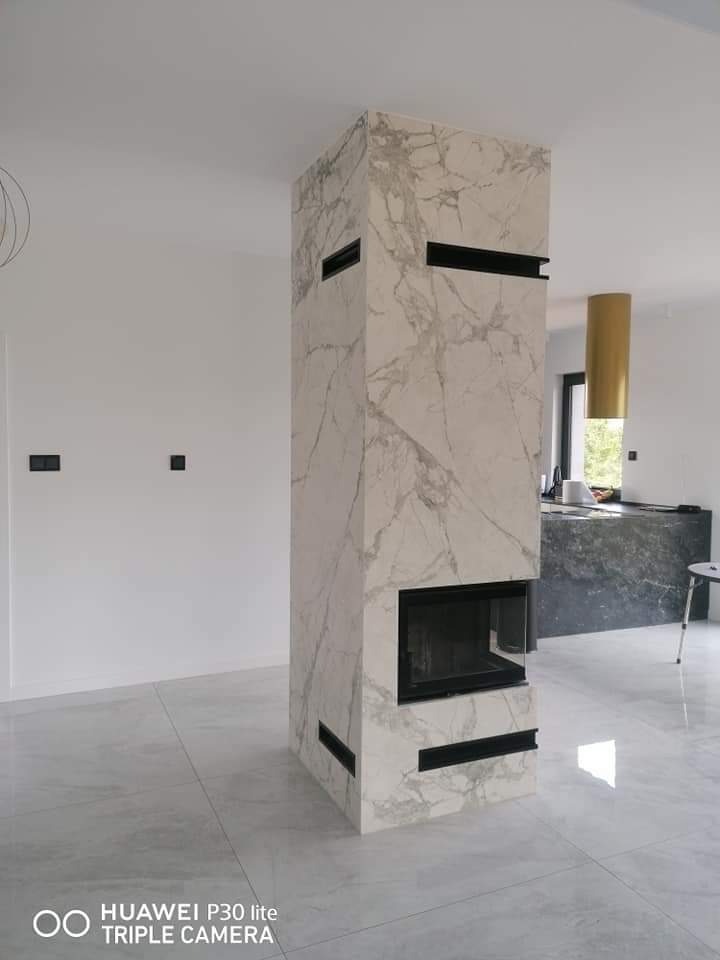 Nowoczesny kominek obudowany płytami imitującymi marmur, z czarnymi wstawkami wentylacyjnymi, na tle jasnej podłogi i ściany.