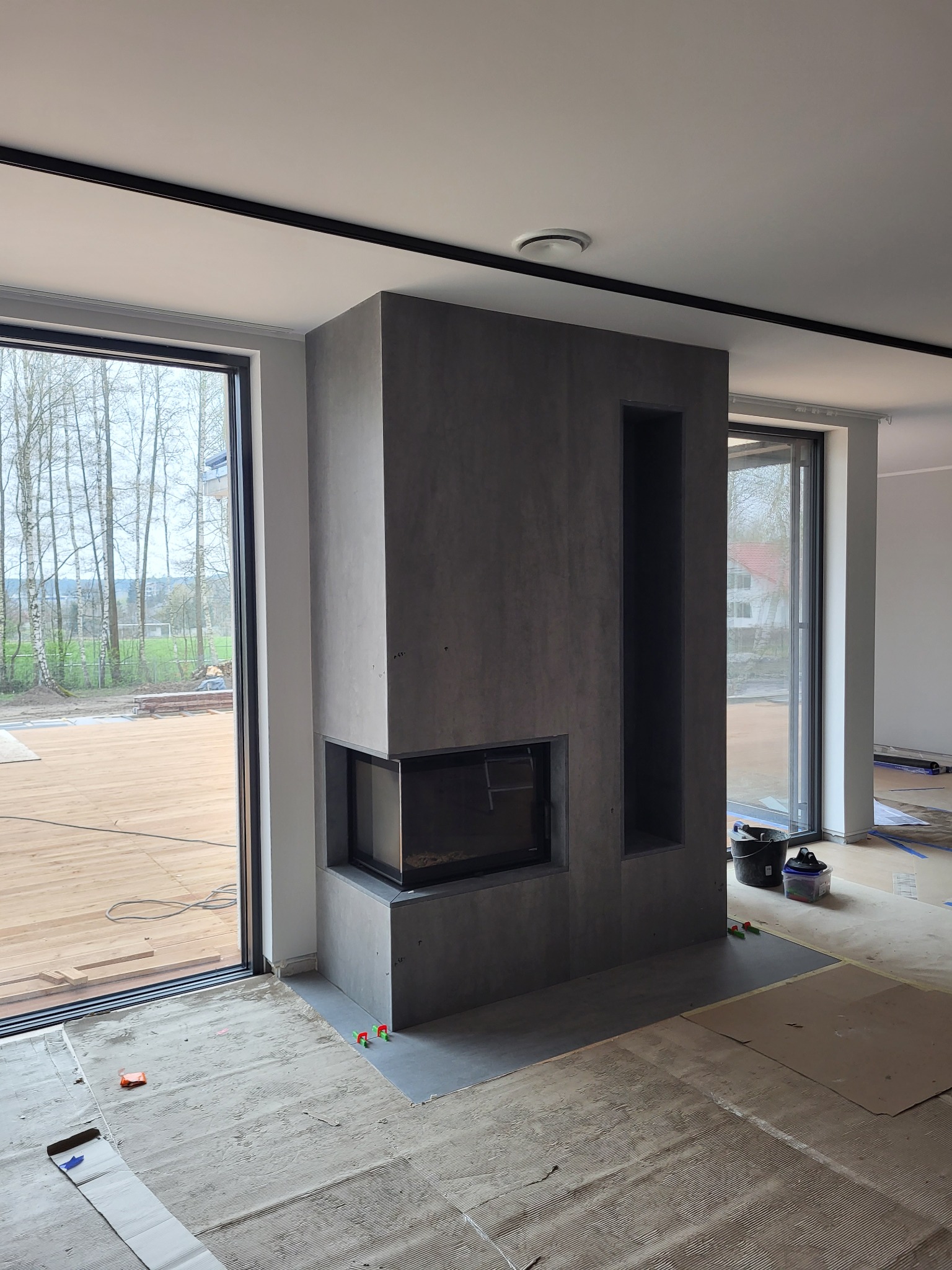 Nowoczesny kominek obudowany szarym kamieniem, z boczną wnęką, w trakcie montażu; podłoga zabezpieczona kartonem, widoczne okna z widokiem na drzewa.