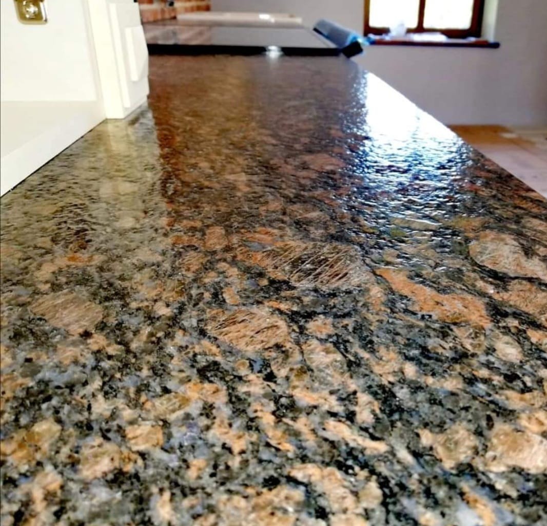 Błyszczący, polerowany blat o nieregularnym wzorze z brązowych, czarnych i szarych plam, widoczna refleksja światła, fragment kuchni w tle.