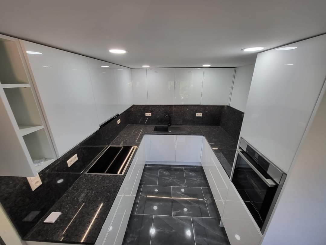 Nowoczesna kuchnia z białymi, lakierowanymi szafkami i ciemnym blatem z wyraźną fakturą kamienia, czarna armatura i płyta indukcyjna, szara podłoga z połyskiem.