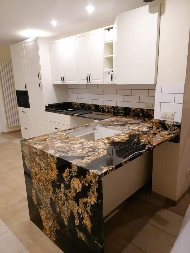 Kuchnia z białymi szafkami i blatem o bogatym, czarno-złotym wzorze kamienia, z widocznym wycięciem na zlew, podświetlona przez punktowe światła sufitowe.