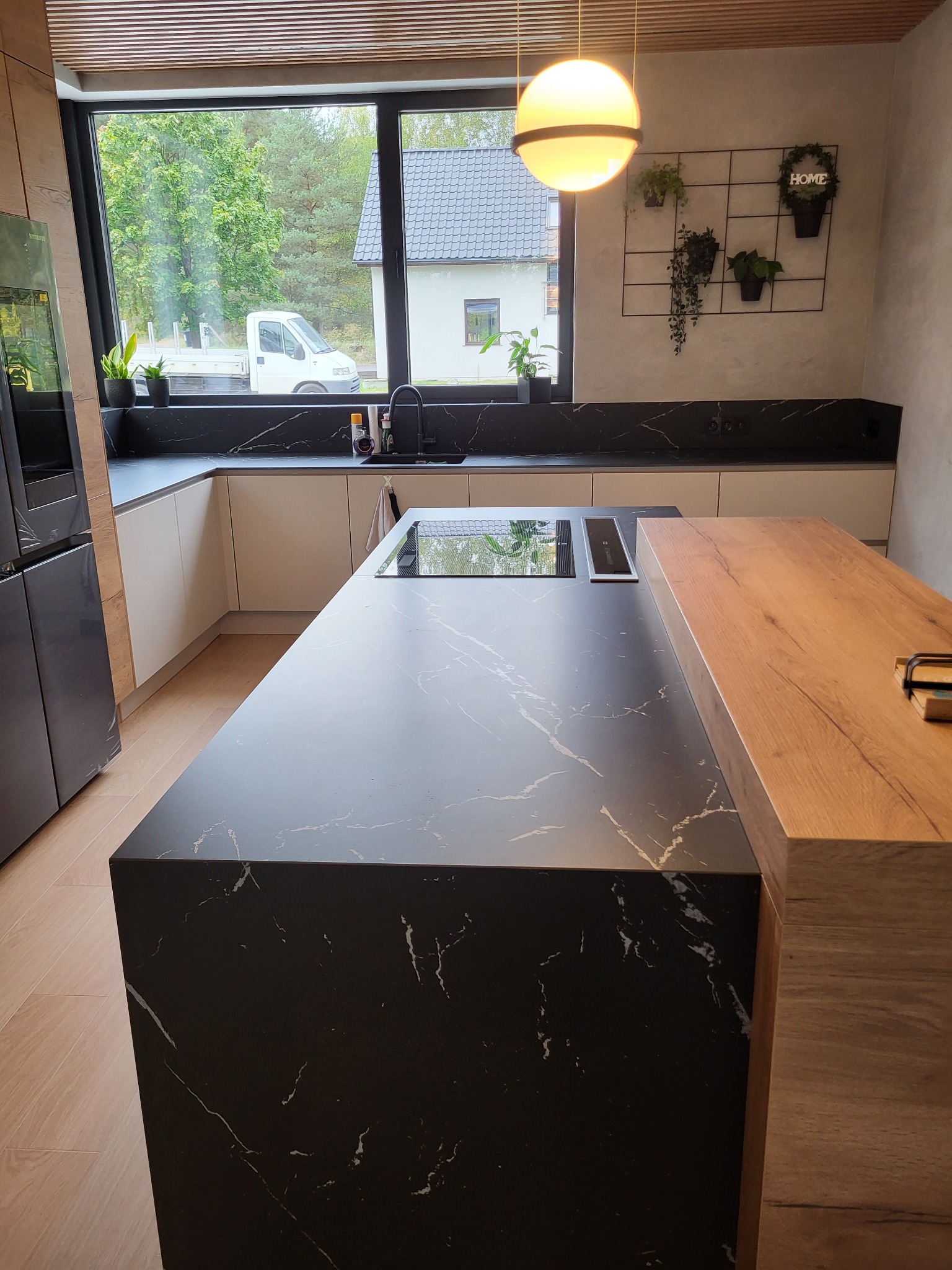 Nowoczesna kuchnia z wyspą i blatem z czarnego kamienia z białymi żyłkami, widok na okno z zielenią i białym samochodem dostawczym na zewnątrz.