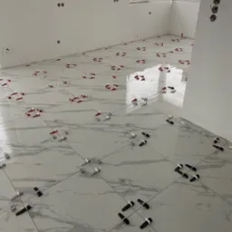Podłoga z płytek imitujących marmur w trakcie układania, z widocznymi krzyżykami dystansowymi w kolorach czerwonym, białym i czarnym, odblask światła na powierzchni płytek, ściany w stanie surowym.