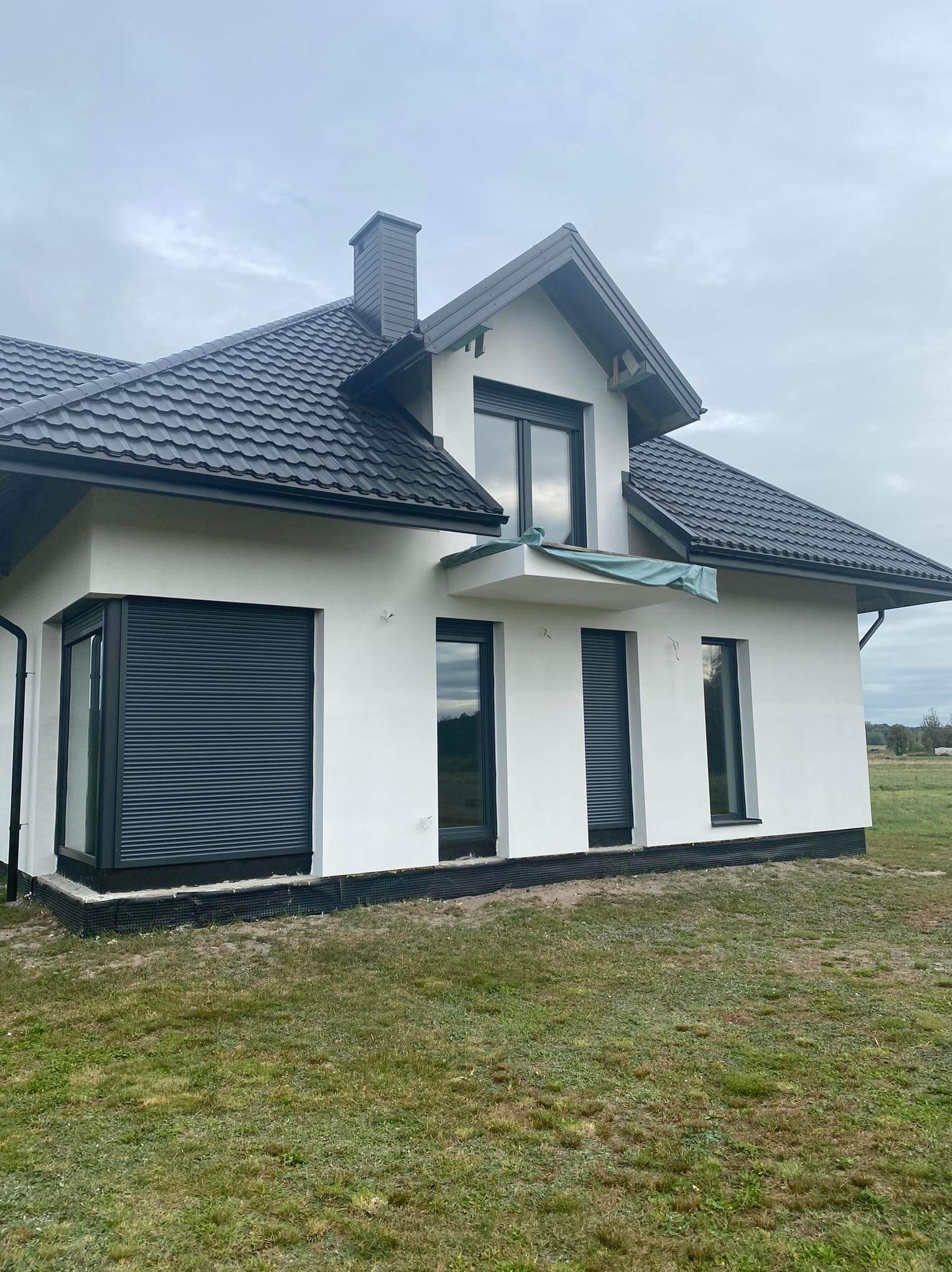 Nowoczesny dom jednorodzinny z czarną dachówką i białymi ścianami, widoczne okna z roletami zewnętrznymi w kolorze antracytowym, częściowo zakryty balkon zieloną plandeką, otoczony trawnikiem.