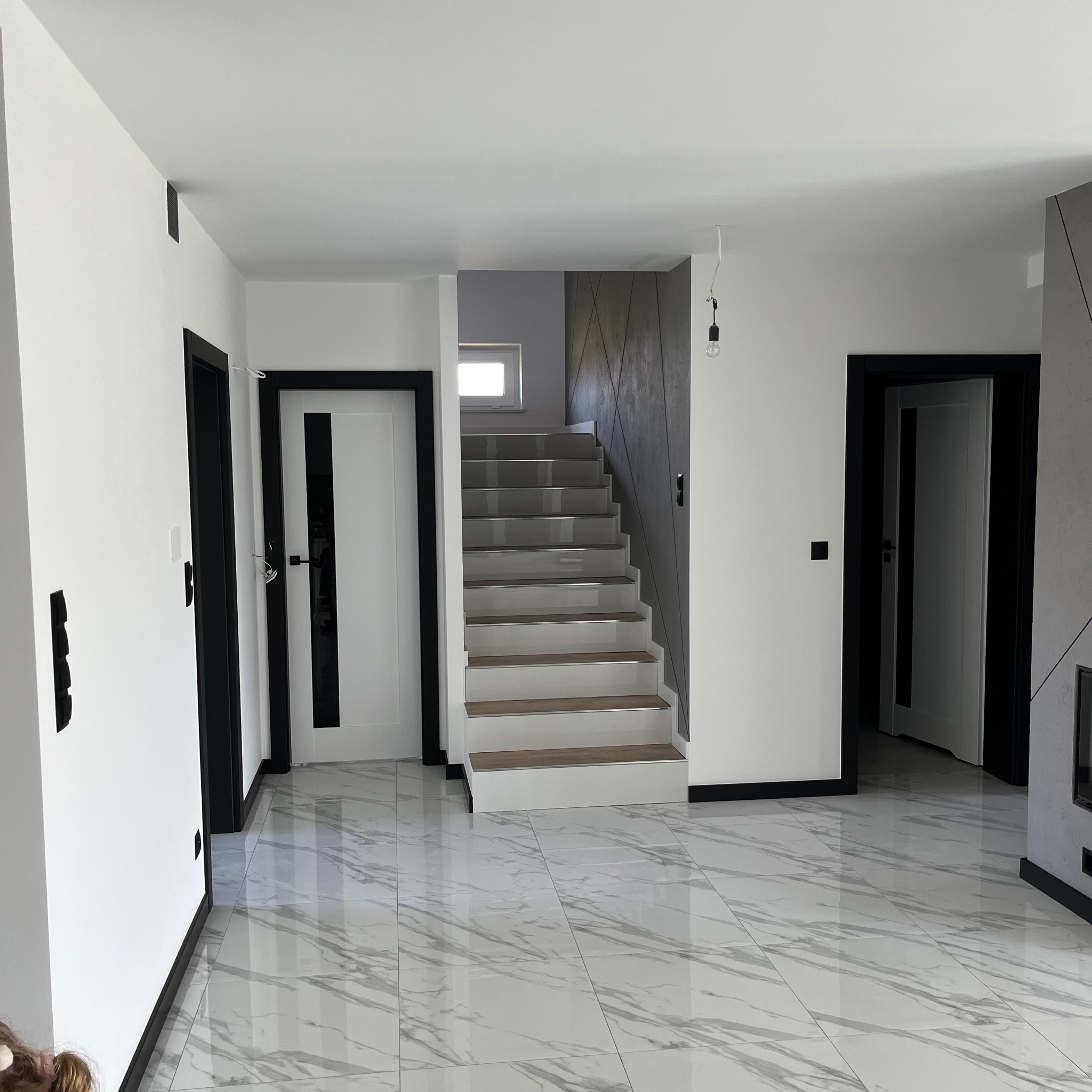 Nowoczesne wnętrze z białymi ścianami, marmurową podłogą i schodami z drewnianymi stopniami, czarne obramowania drzwi, surowa żarówka zwisająca z sufitu.