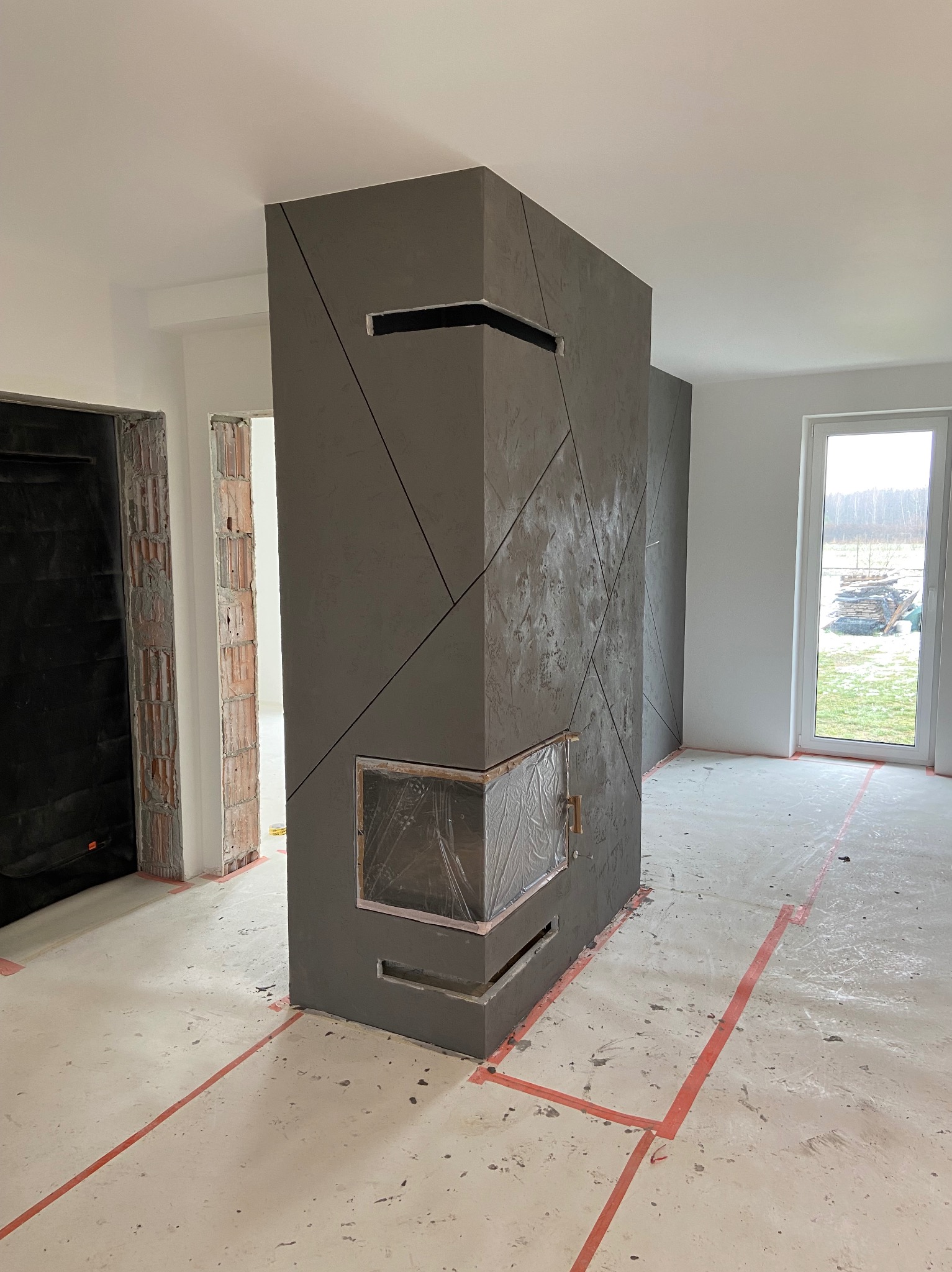 Nowoczesny, ciemnoszary kominek obłożony dekoracyjnym tynkiem strukturalnym z geometrycznym wzorem, zabezpieczony folią, w surowym wnętrzu z widocznymi śladami prac wykończeniowych i oznaczeniami...