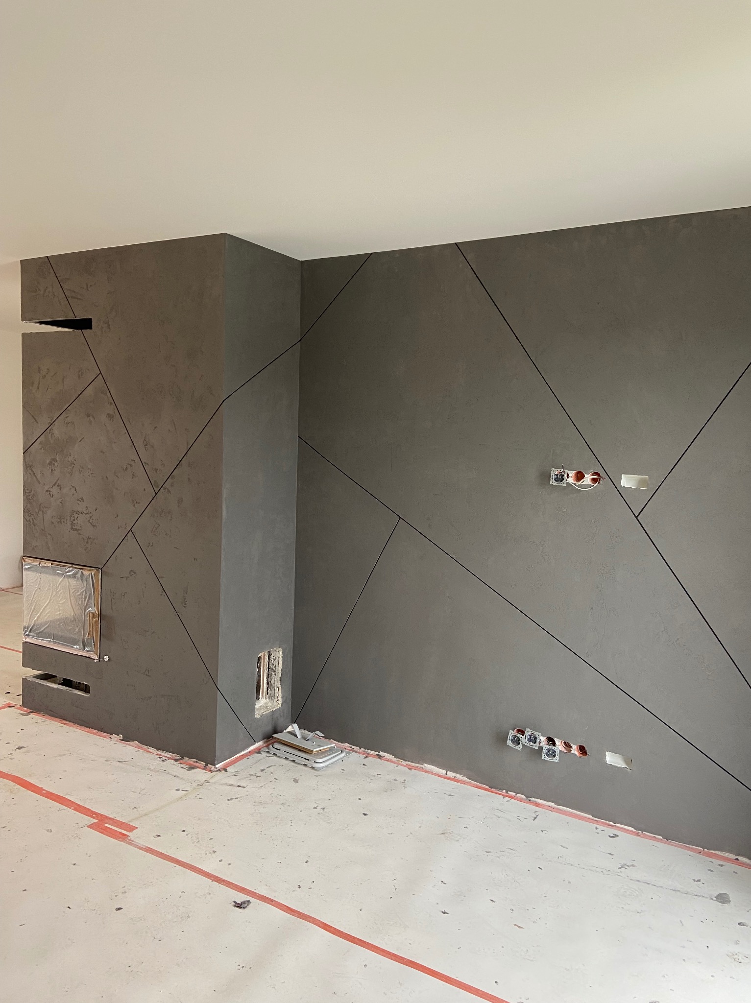 Szara ściana z dekoracyjną gładzią w geometryczne wzory, widoczne puszki elektryczne i zabezpieczony folią kominek, podłoga z resztkami po remoncie i czerwoną taśmą wyznaczającą obszar.