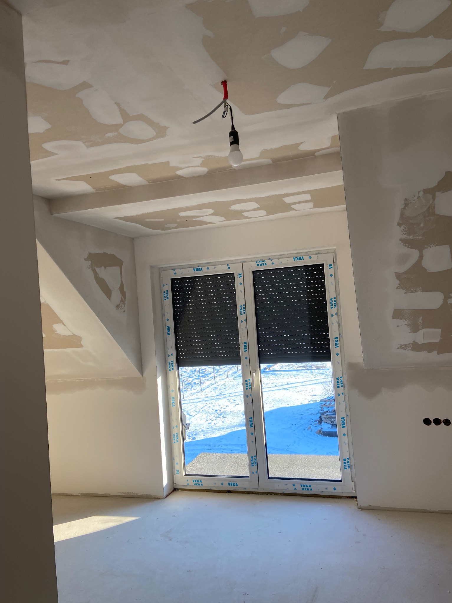 Pokój w trakcie prac wykończeniowych: ściany i sufit z widocznymi łatami świeżo nałożonej masy szpachlowej, okno z roletami zewnętrznymi, surowa żarówka na kablu zwisająca z sufitu.