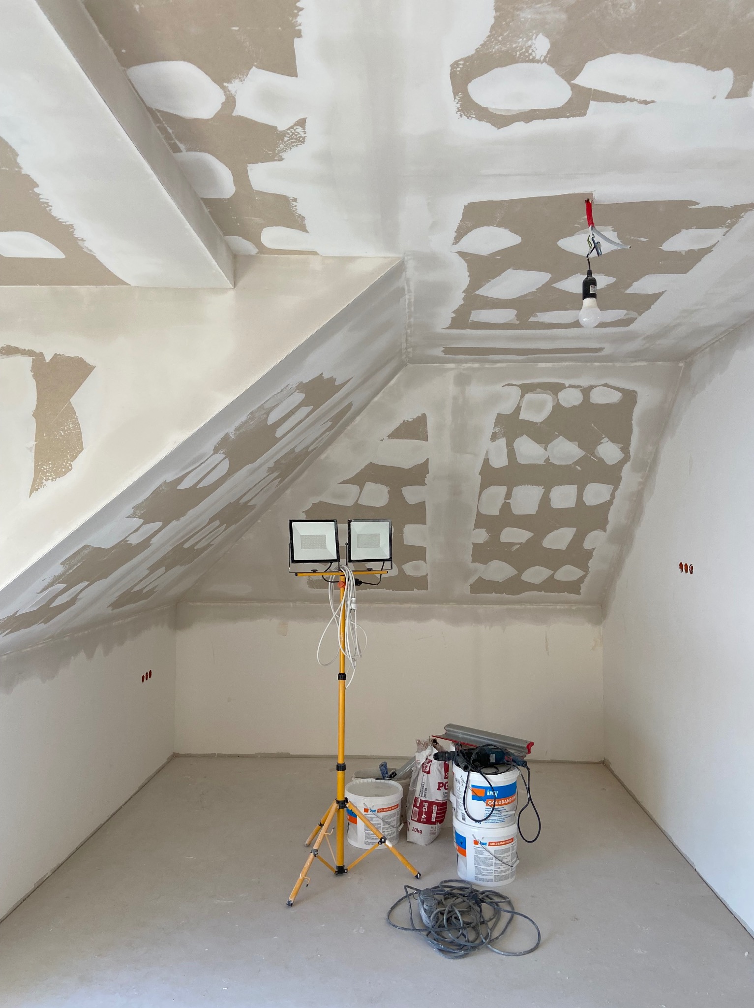 Poddasze w trakcie prac wykończeniowych, widoczne ślady po łataniu sufitu i ścian, żółty statyw z dwoma reflektorami, wiadra z masą szpachlową i narzędzia na podłodze.