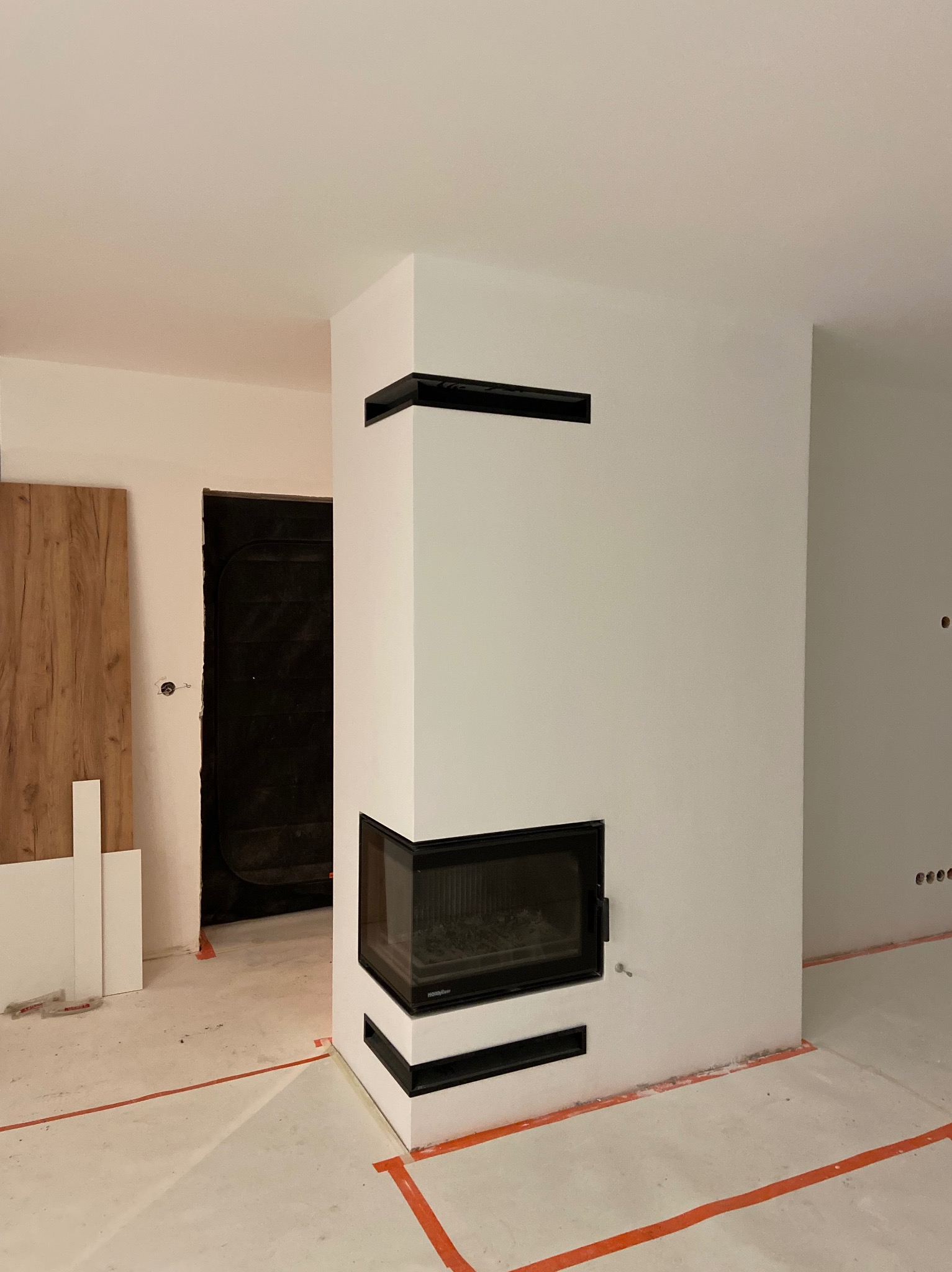 Nowoczesny, narożny kominek z czarnymi akcentami w trakcie wykończenia, widoczne taśmy malarskie na podłodze i niedokończone ściany.