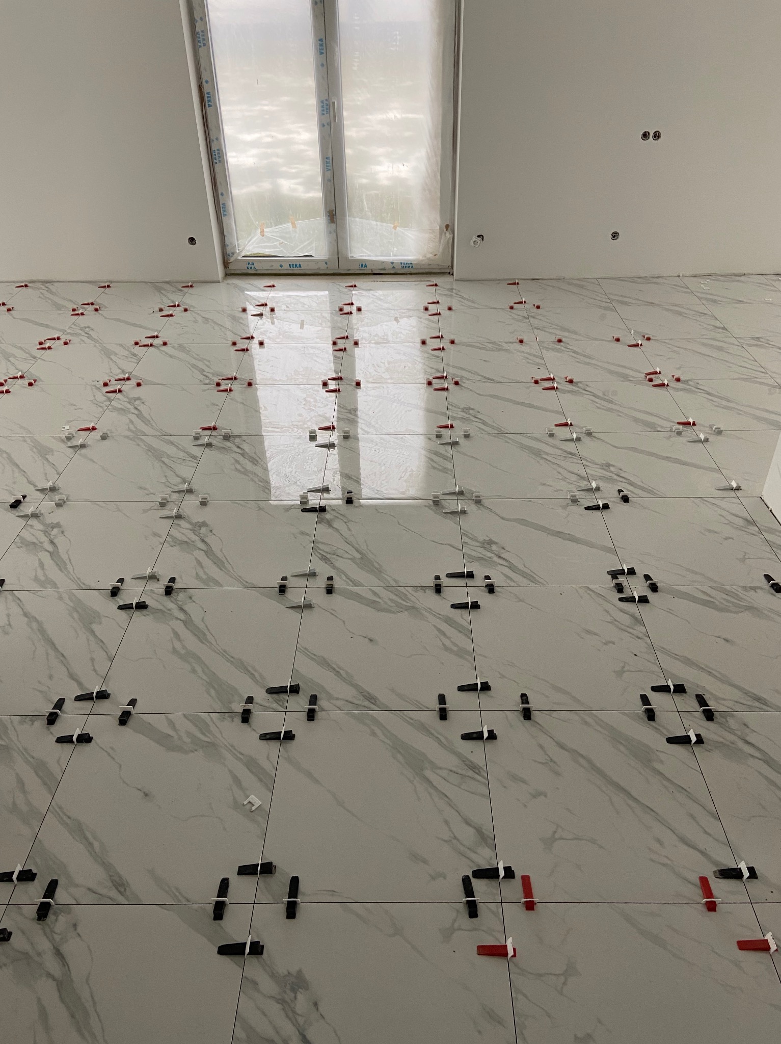 Świeżo ułożone, białe płytki imitujące marmur z czerwonymi, białymi i czarnymi klipsami poziomującymi, widoczne otwory na gniazdka elektryczne w ścianie i okno w górnej części kadru.