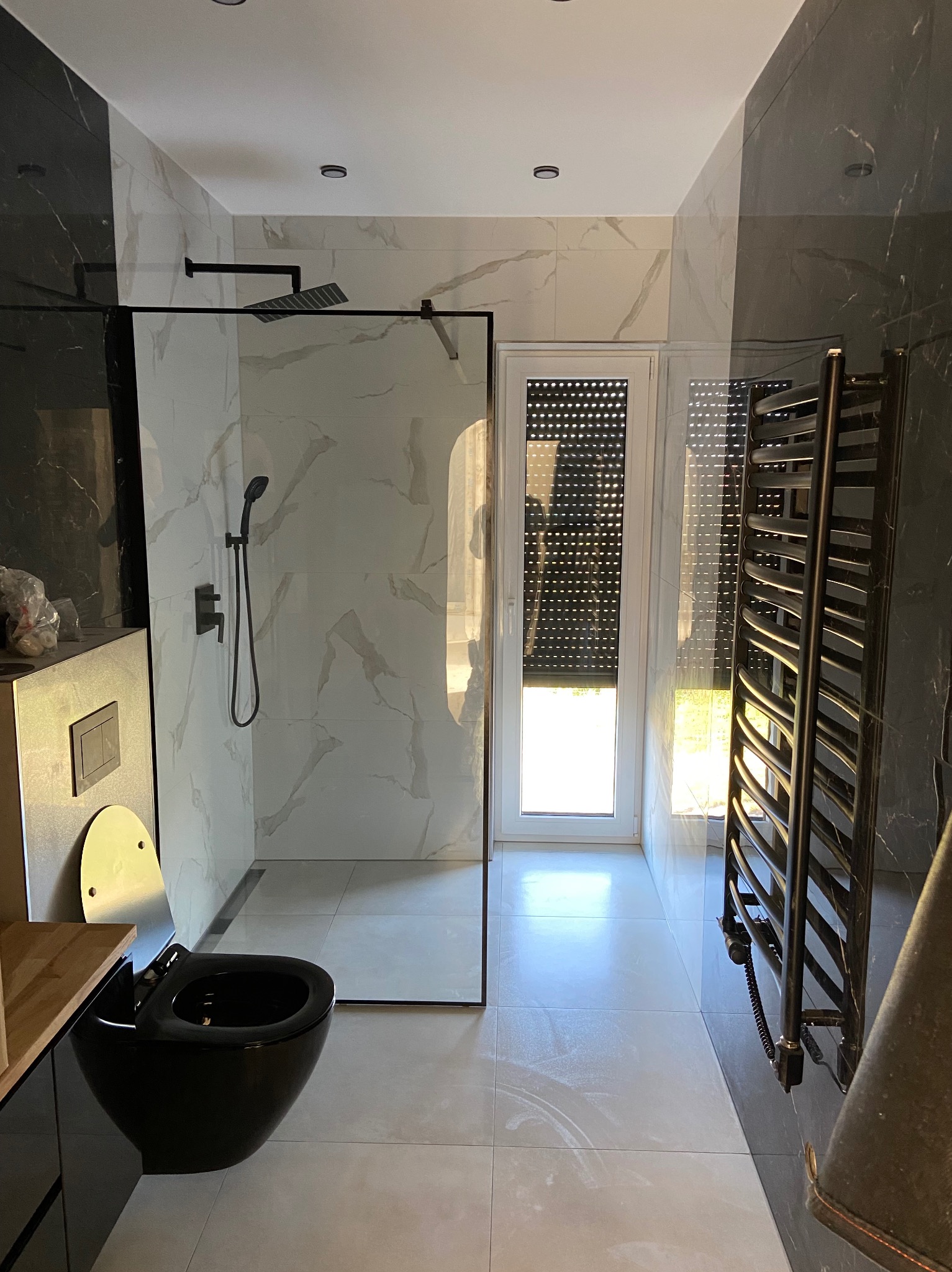 Nowoczesna łazienka z czarną armaturą, kabiną prysznicową typu walk-in z białymi płytkami imitującymi marmur, wiszącą toaletą i grzejnikiem drabinkowym, widok na okno z roletami.