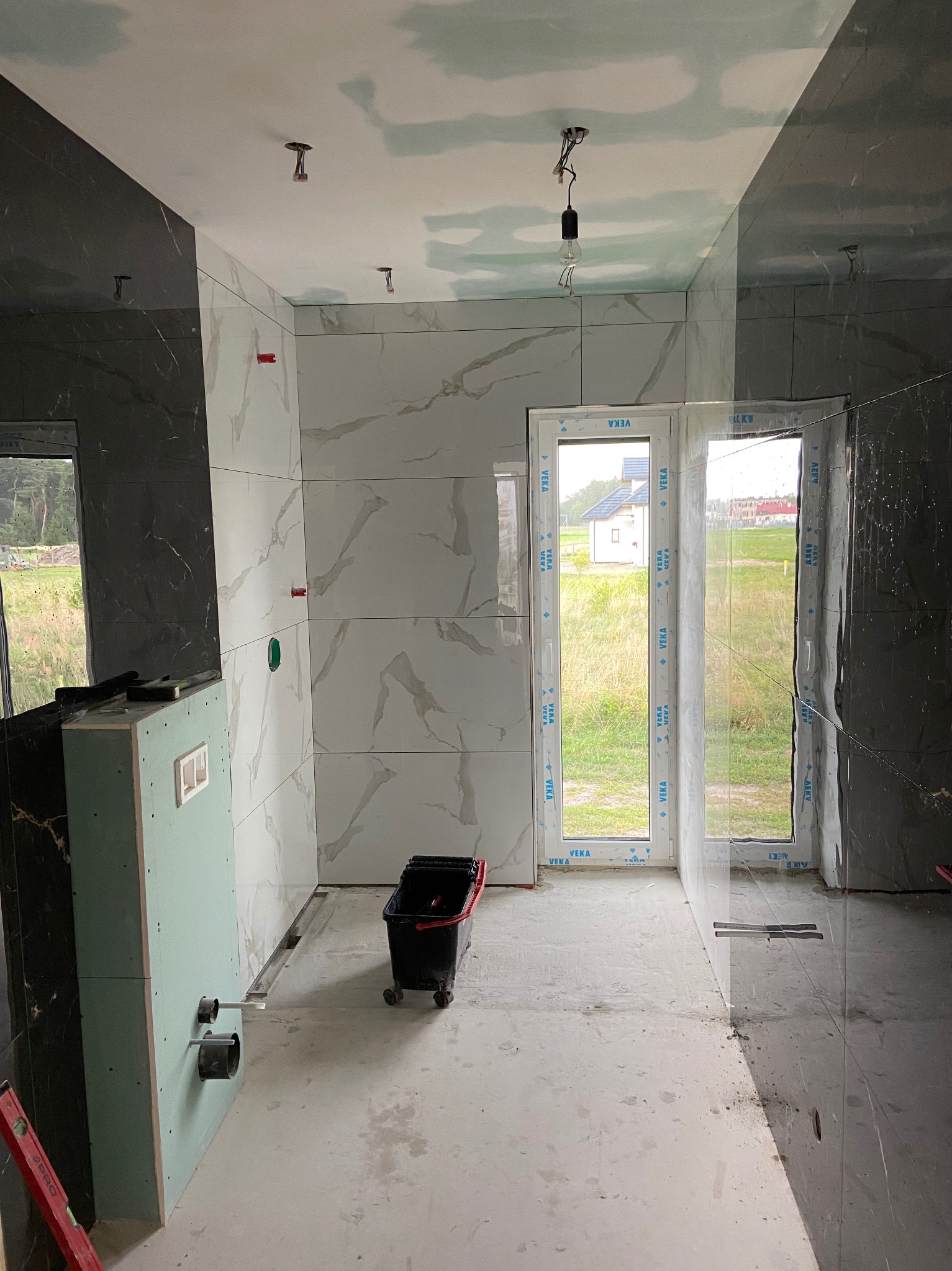 Wnętrze łazienki w trakcie remontu: częściowo wyłożone płytkami imitującymi marmur w odcieniach szarości, z widoczną konstrukcją podtynkową z płyty karton gips, surowy beton na podłodze, okno...