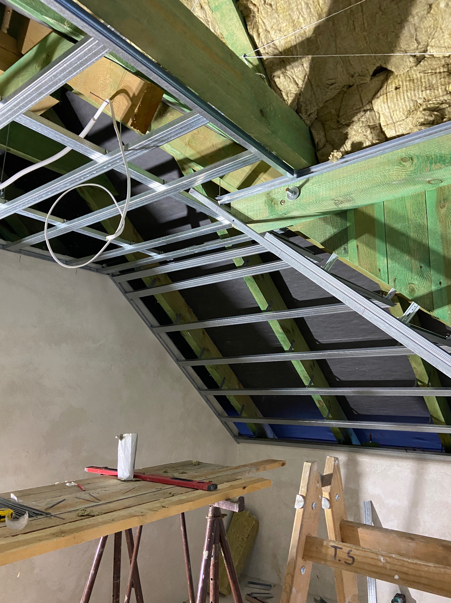Metalowa konstrukcja sufitu podwieszanego z widoczną izolacją na poddaszu, ustawiona pod kątem, stolik roboczy z poziomnicą i narzędziami oraz drewniana drabina w tle.