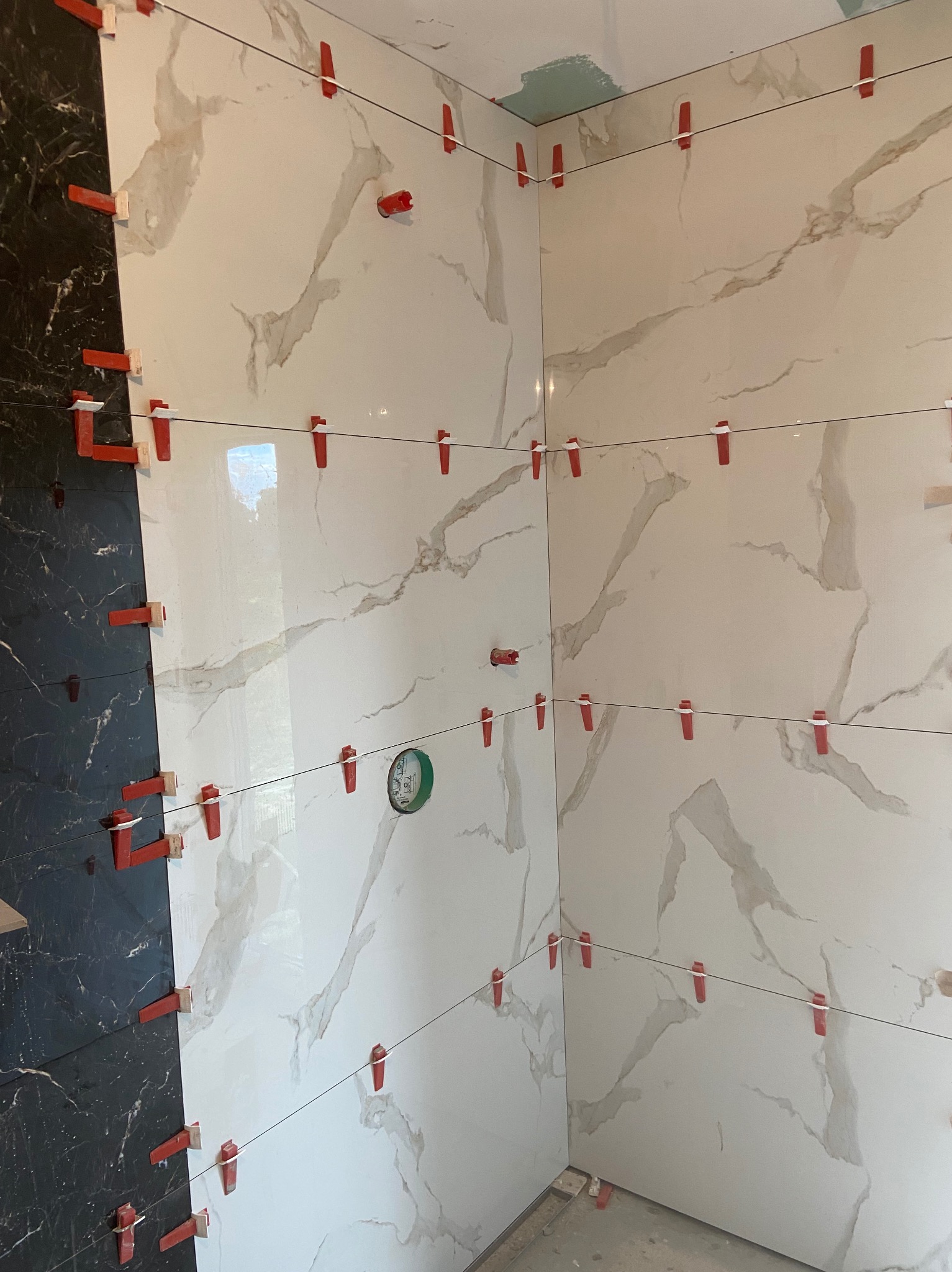 Montaż płytek w narożniku łazienki, widoczne czarne i białe płytki z marmurkowym wzorem, system poziomowania płytek z czerwonymi klipsami i żyłką, puszka elektryczna w ścianie.