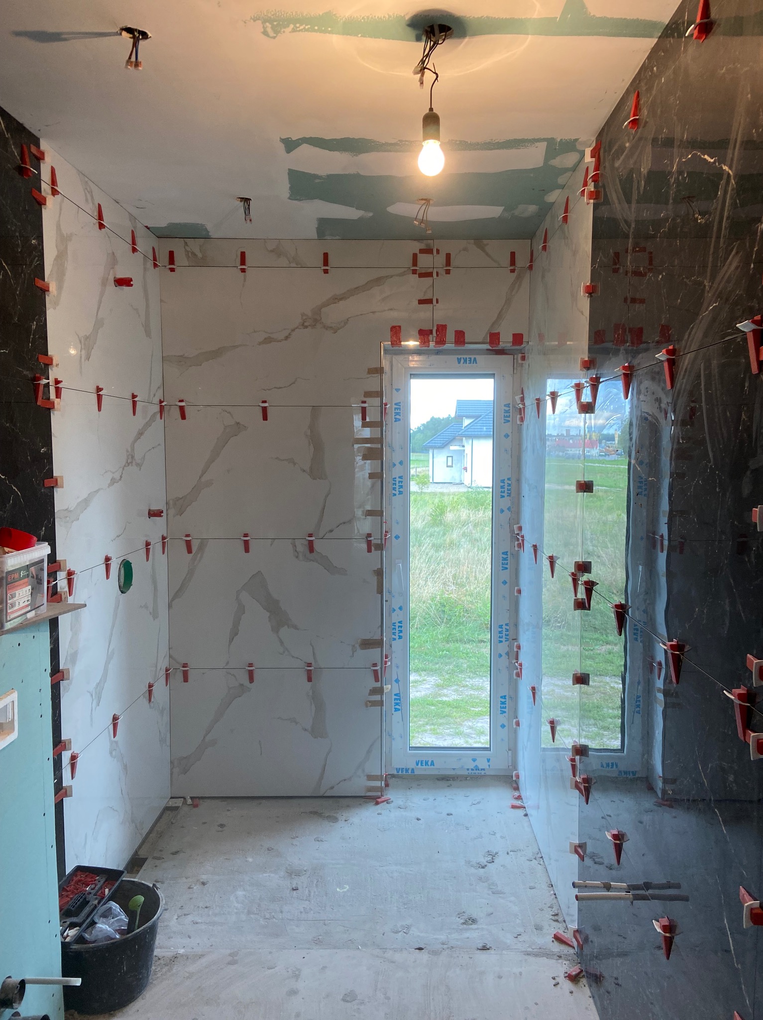 Łazienka w trakcie remontu, z częściowo ułożonymi płytkami imitującymi marmur, czarne płytki na ścianie po prawej, widoczne czerwone klipsy poziomujące, okno z widokiem na dom i trawnik, surowy...