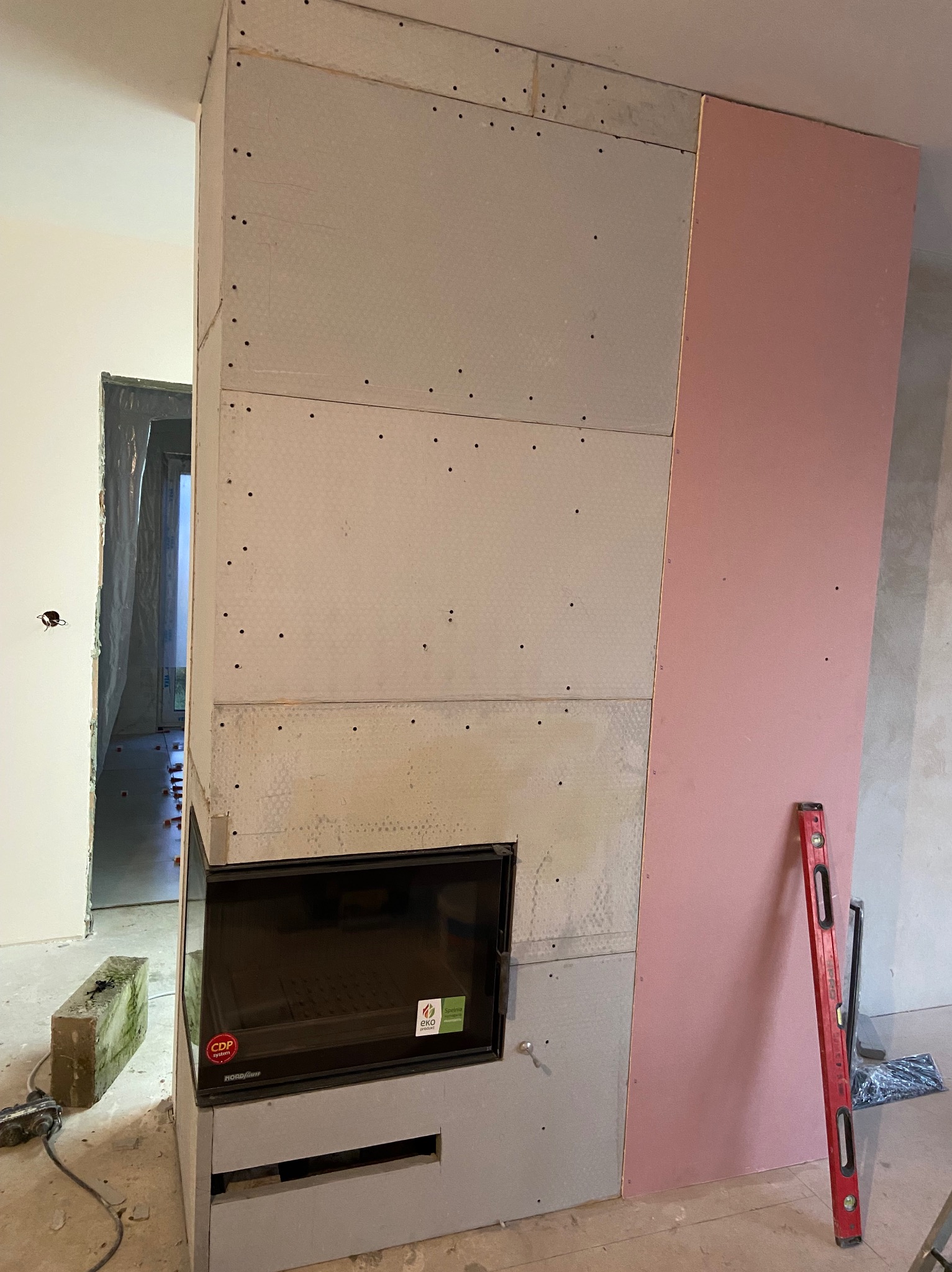 Wnętrze w trakcie prac wykończeniowych: zabudowa kominka z płyt kartonowo-gipsowych, widoczny wkład kominkowy, poziomica oparta o ścianę, fragment podłogi z kaflami i folią ochronną.