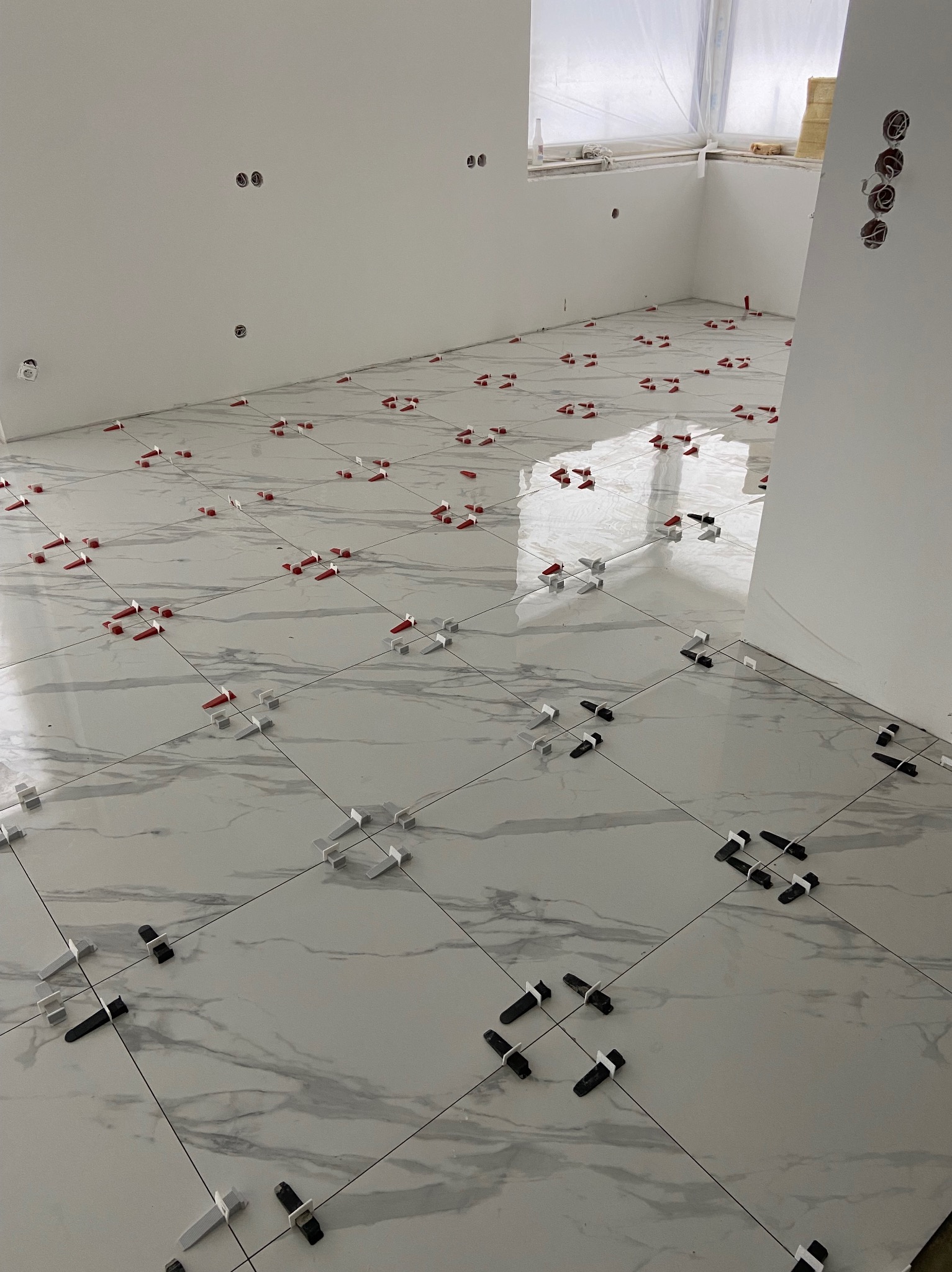 Podłoga z płytek imitujących marmur w trakcie układania, z widocznymi krzyżykami dystansowymi w kolorach czerwonym, białym i czarnym, odblask światła na powierzchni płytek, ściany w stanie surowym.