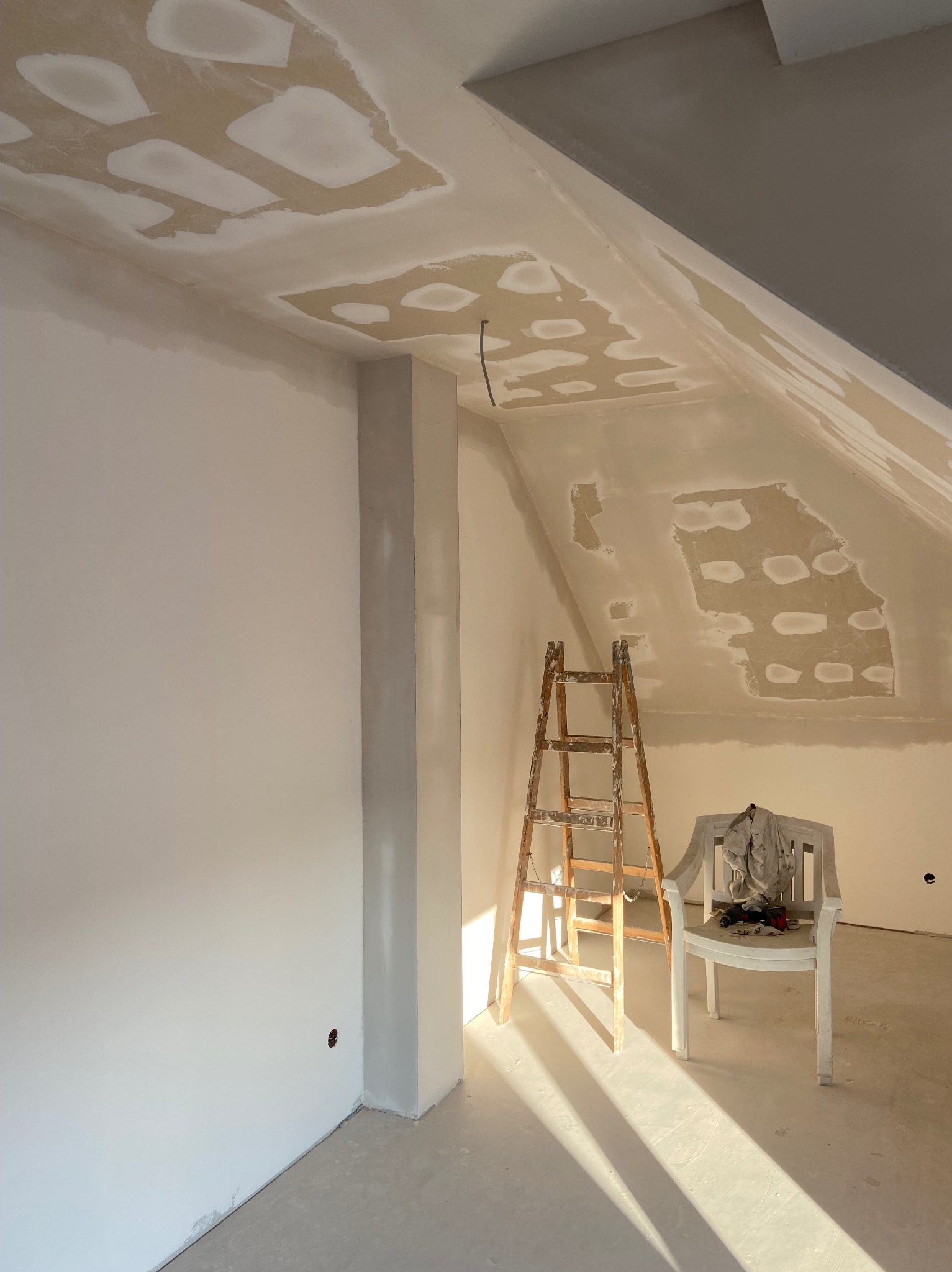 Pokój w trakcie remontu: ściany i sufit po łataniu płyt kartonowo-gipsowych, widoczna drabina oparta o ścianę, na krześle leżą narzędzia i ubranie robocze.