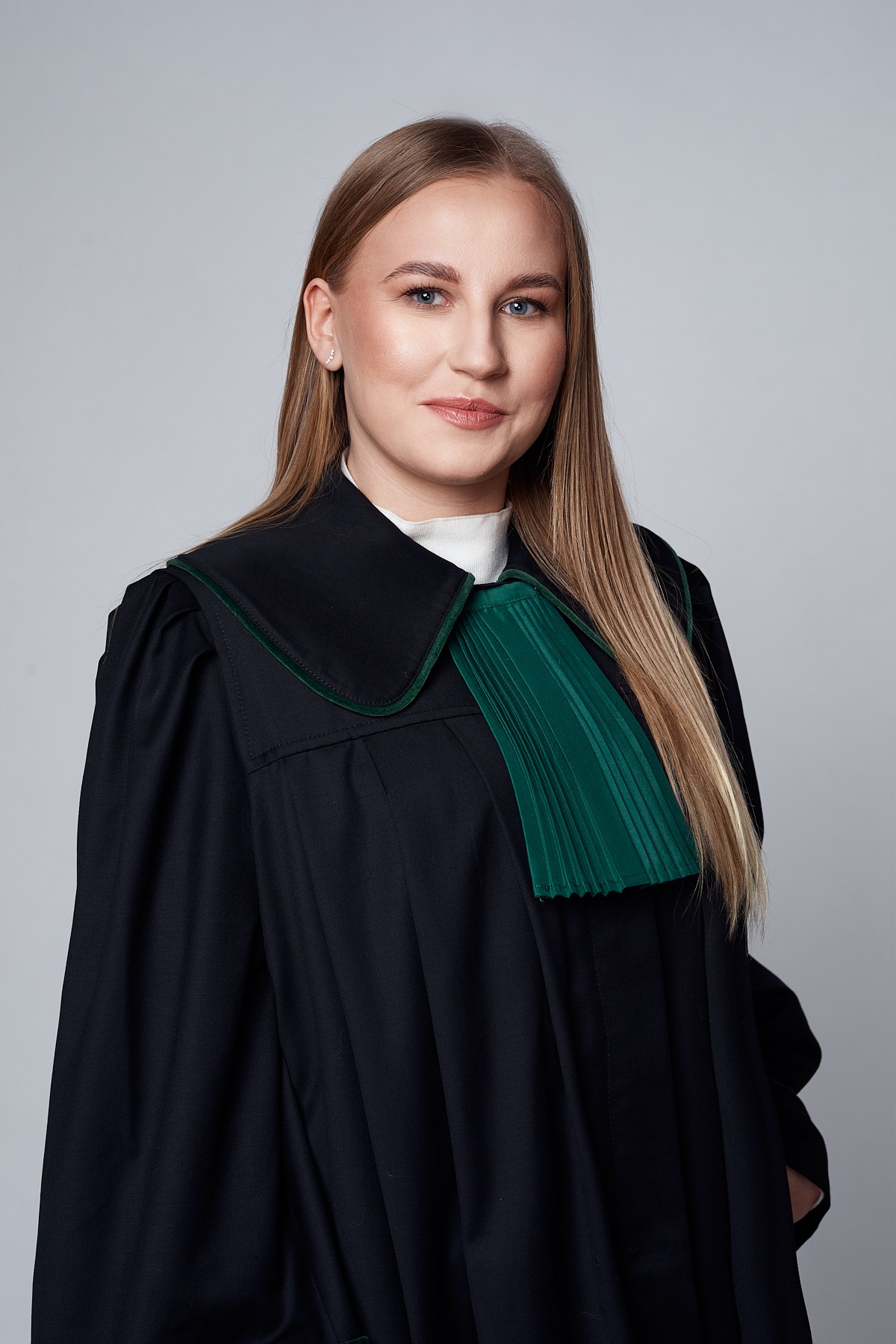 Portret młodej kobiety z blond włosami, ubranej w togę adwokacką z zielonym żabotem, na jednolitym szarym tle.