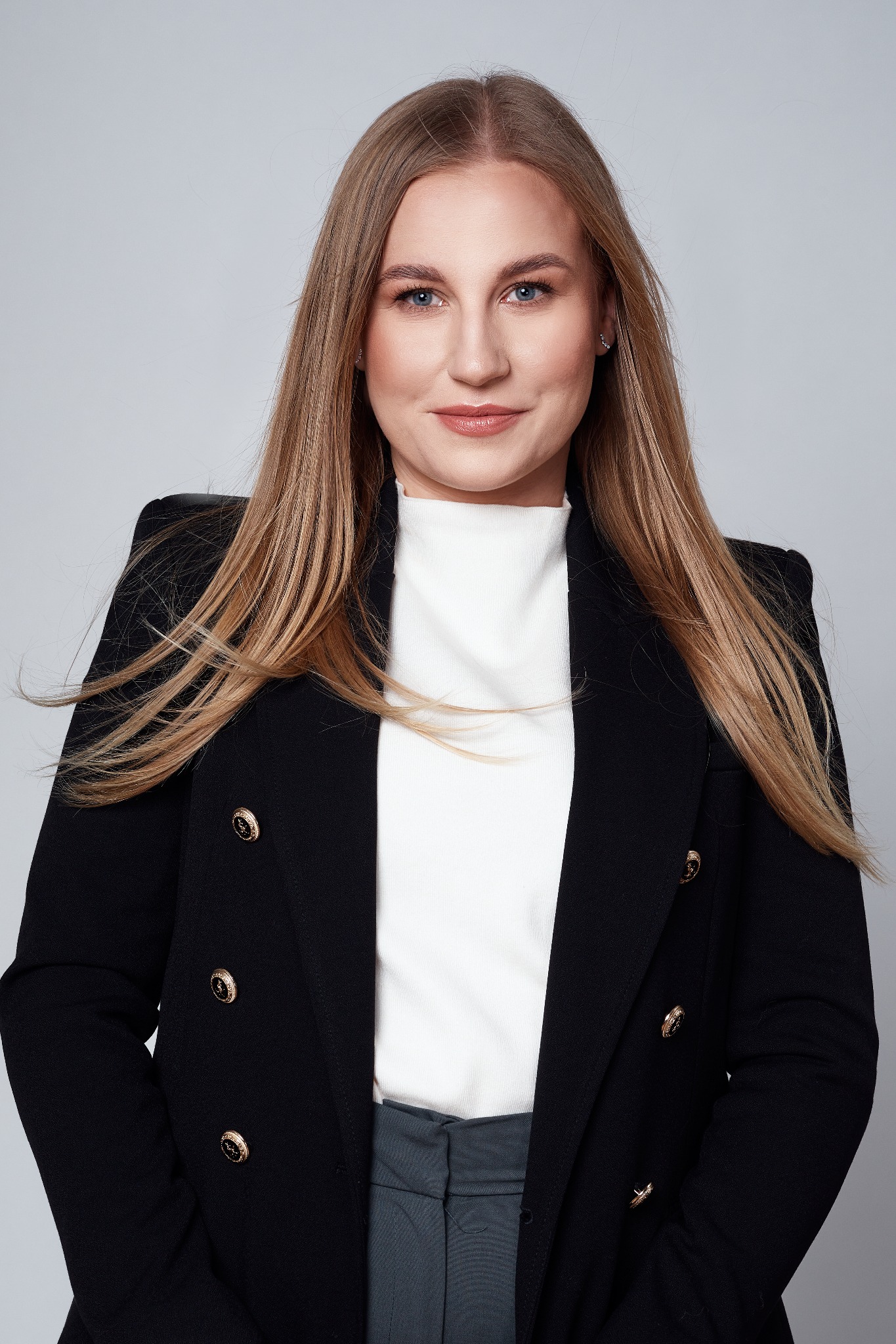 Portret kobiety w czarnej marynarce z ozdobnymi złotymi guzikami i białym golfie na jednolitym szarym tle.