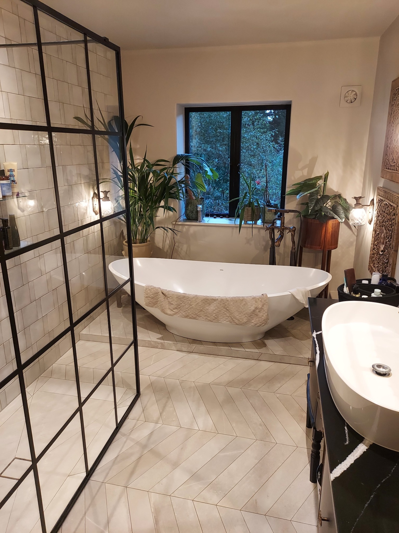 Nowoczesna łazienka z wolnostojącą wanną, kabiną prysznicową typu walk-in z czarnymi profilami, umywalką nablatową i podłogą z płytek ułożonych w jodełkę; widok na zieleń za oknem.