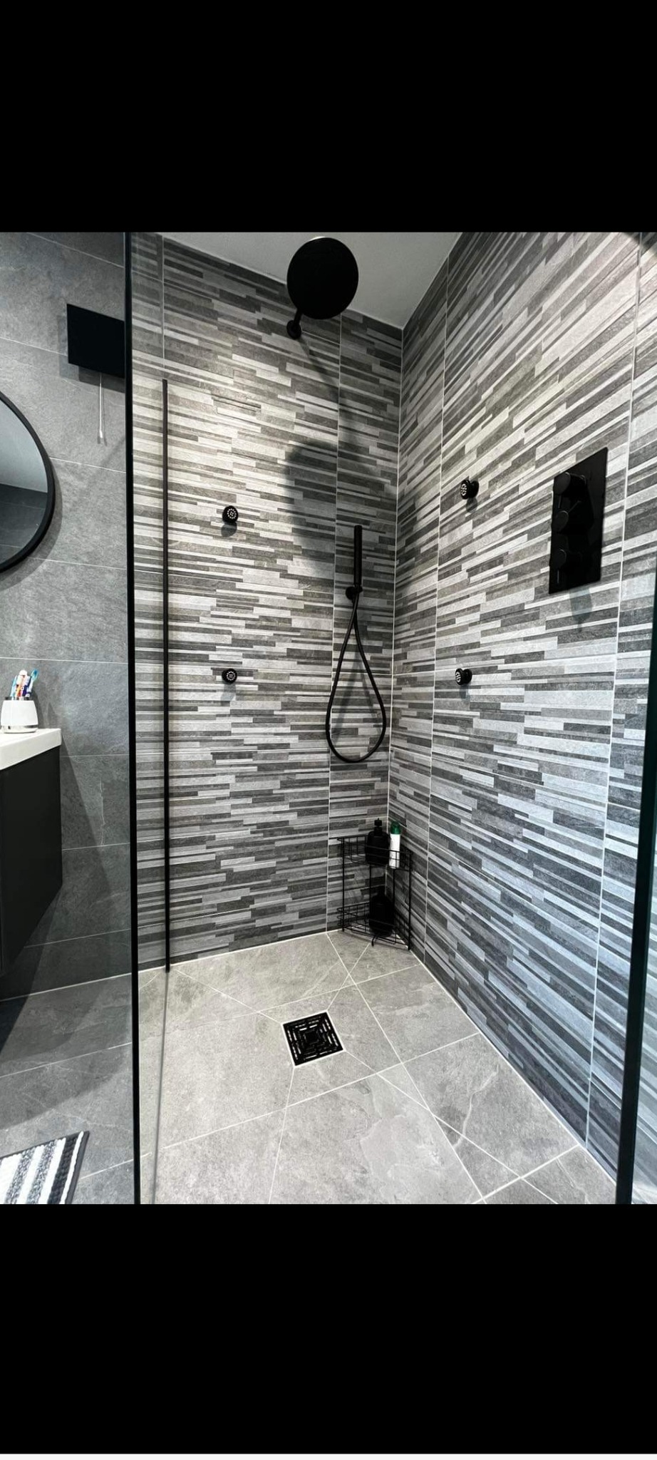 Nowoczesna kabina prysznicowa z szarymi płytkami imitującymi drewno, czarna armatura i odpływ liniowy w podłodze wyłożonej szarymi płytkami.