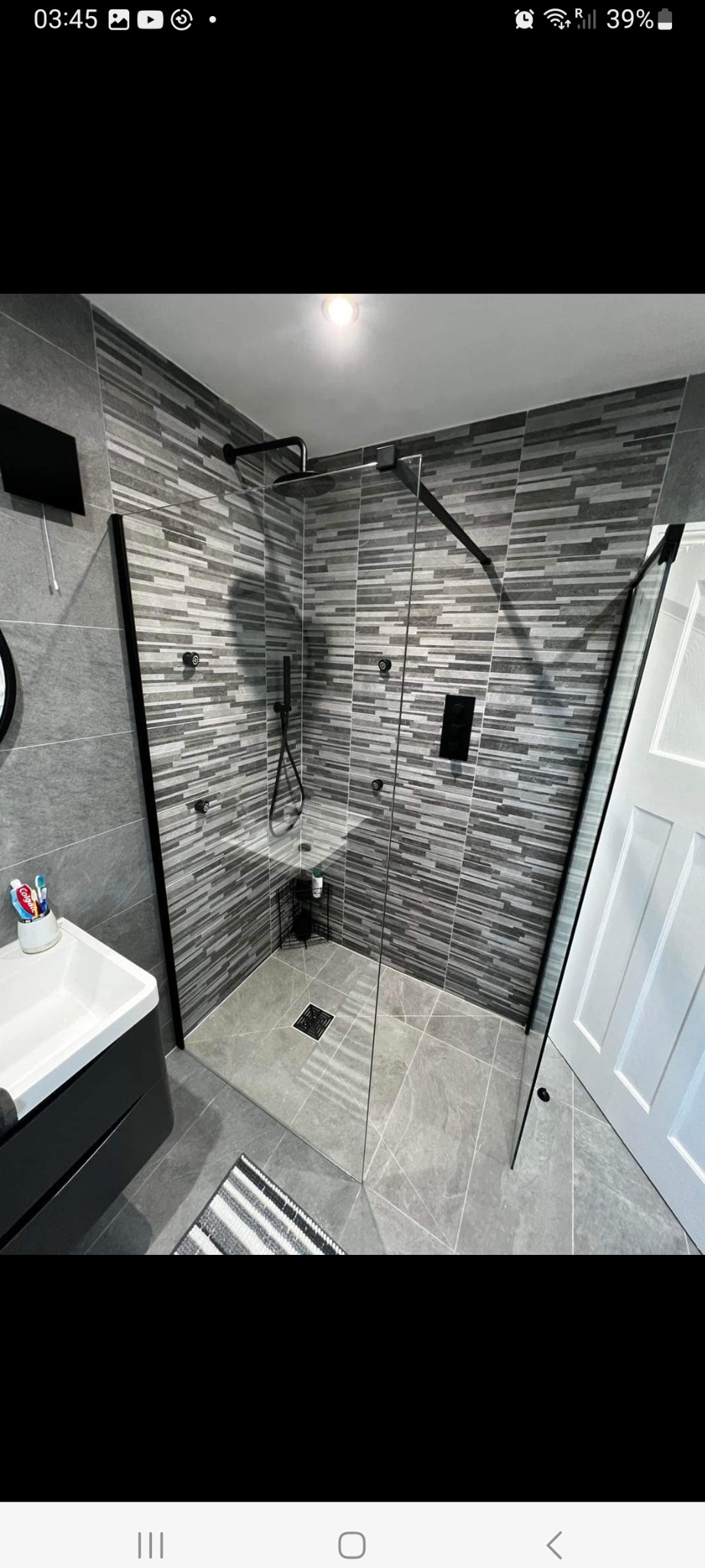 Nowoczesna kabina prysznicowa z czarnymi elementami wykończenia, szare płytki imitujące kamień na ścianach, szara podłoga z odpływem liniowym, widoczna umywalka z czarną szafką i białą misą,...