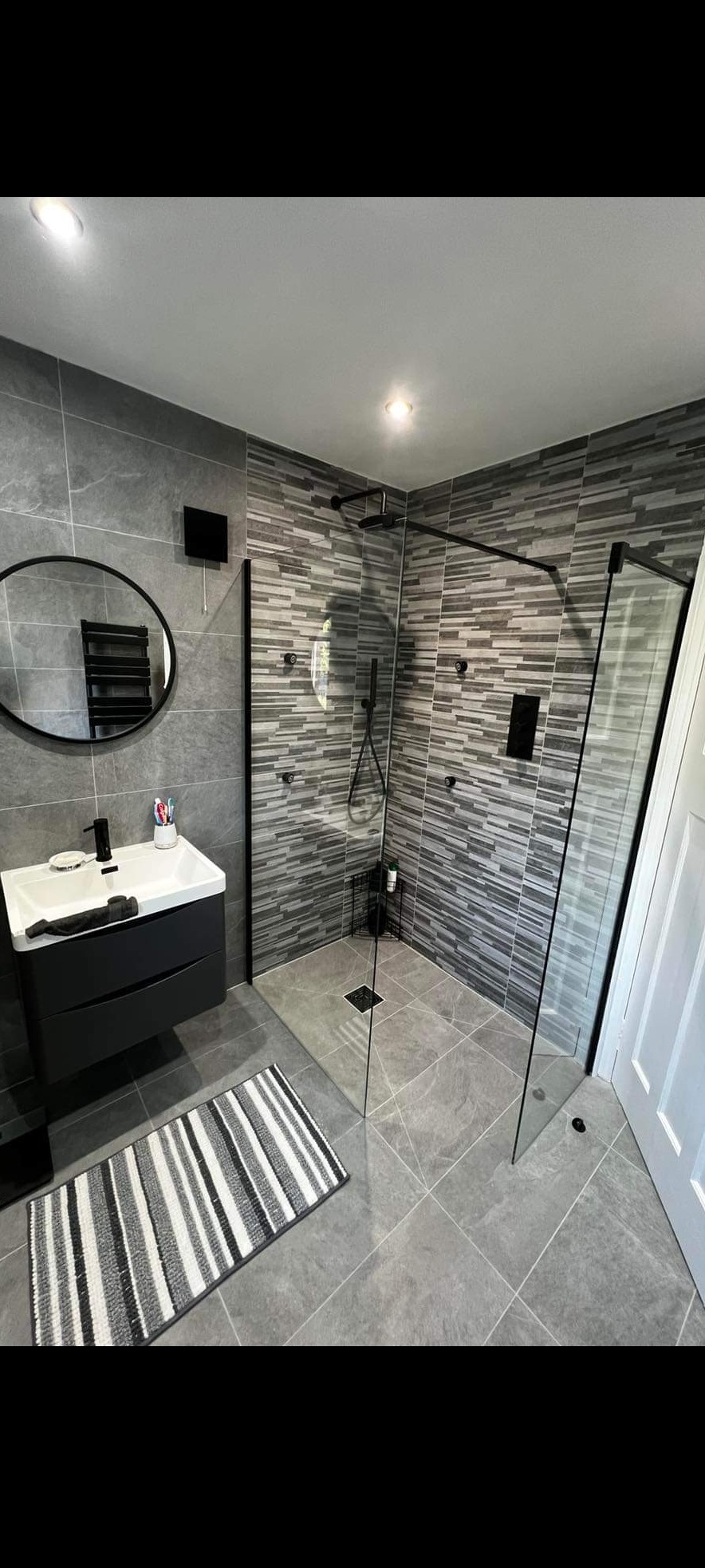 Nowoczesna łazienka z szarymi płytkami, kabiną prysznicową typu walk-in z czarnymi profilami, czarną szafką pod umywalką i okrągłym lustrem z grzejnikiem drabinkowym.