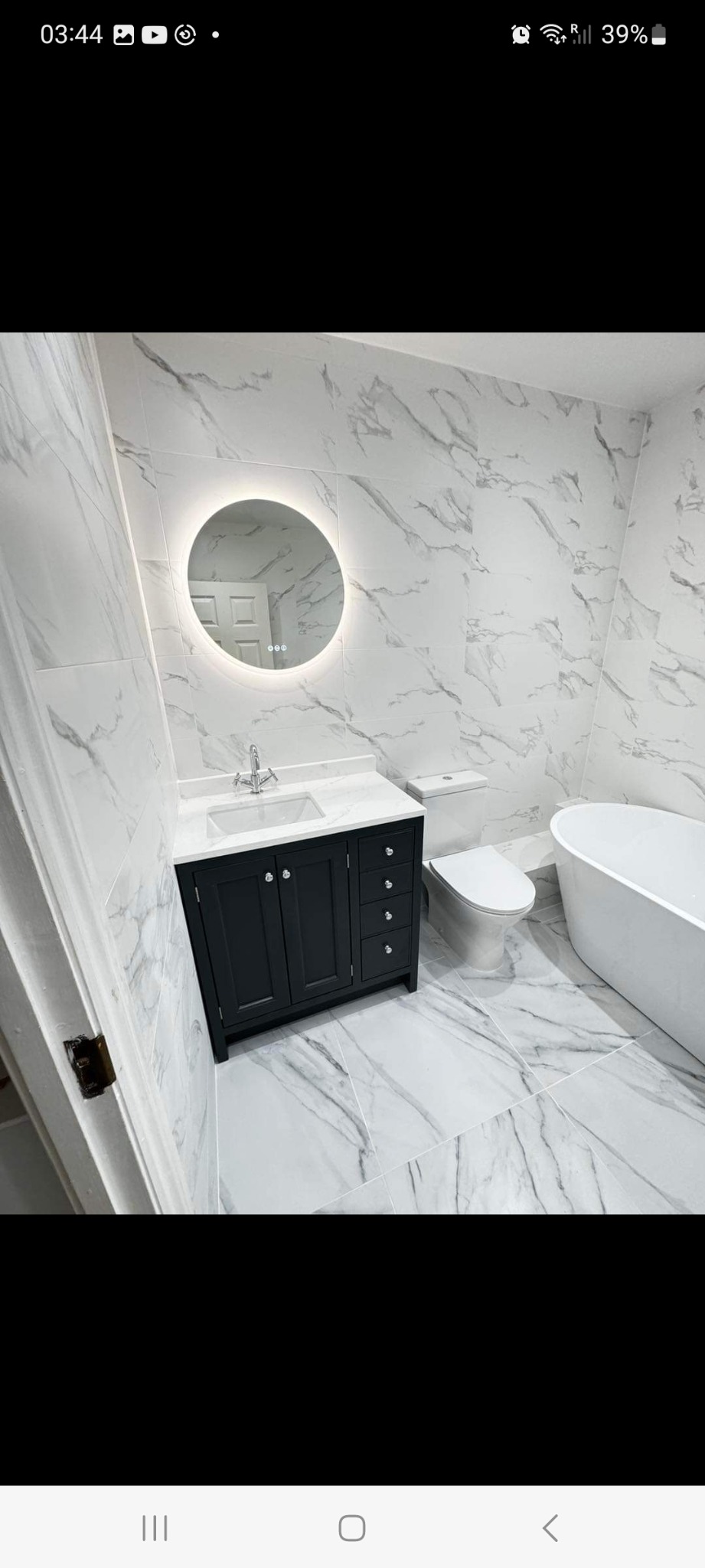 Wykończona łazienka z białymi płytkami imitującymi marmur na ścianach i podłodze, czarna szafka pod umywalką z marmurowym blatem, okrągłe lustro z podświetleniem LED, biała toaleta i wanna...