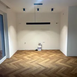 Pusty pokój z ułożonymi panelami w jodełkę, świeżo pomalowanymi ścianami, nowoczesnym oświetleniem sufitowym i widocznym wiadrem z farbą w rogu.