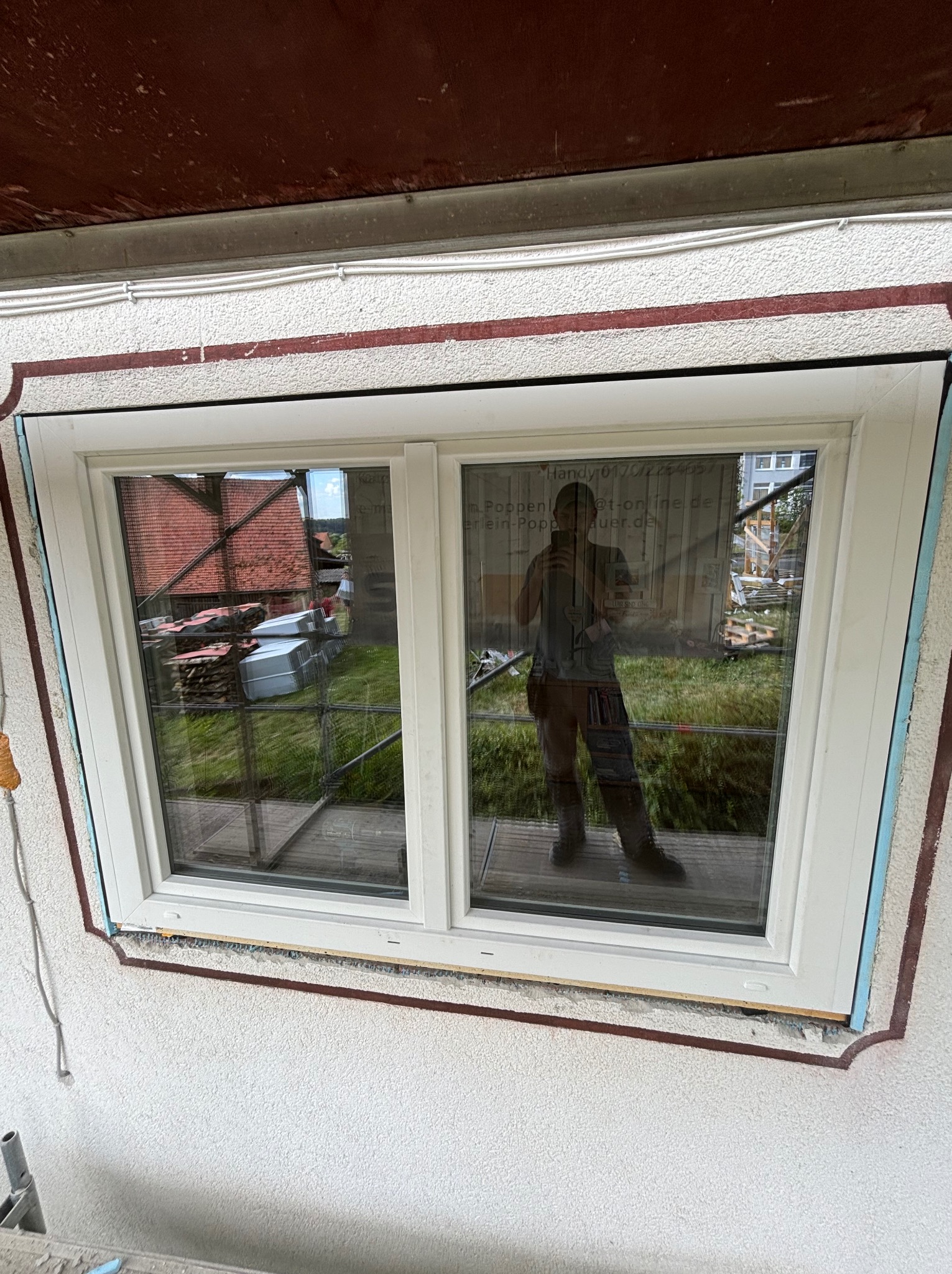 Nowe okno PCV z podwójnymi szybami zamontowane w elewacji budynku, widoczne ślady pianki montażowej i odbicie fotografa w szybie.