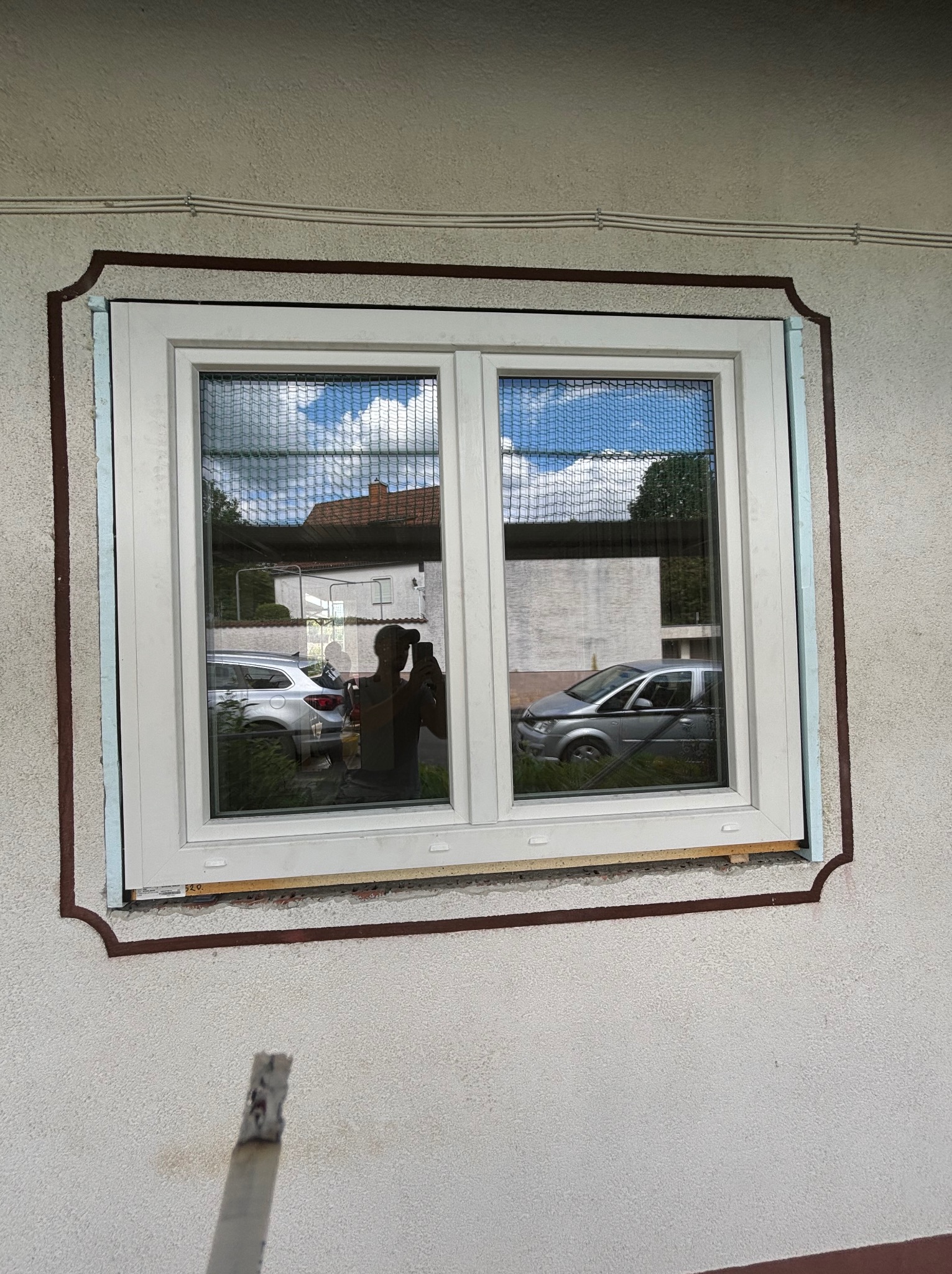 Nowe okno z siatką w białej ramie, widoczna pianka montażowa i odbicie otoczenia w szybach, na elewacji budynku z ozdobnym obramowaniem.