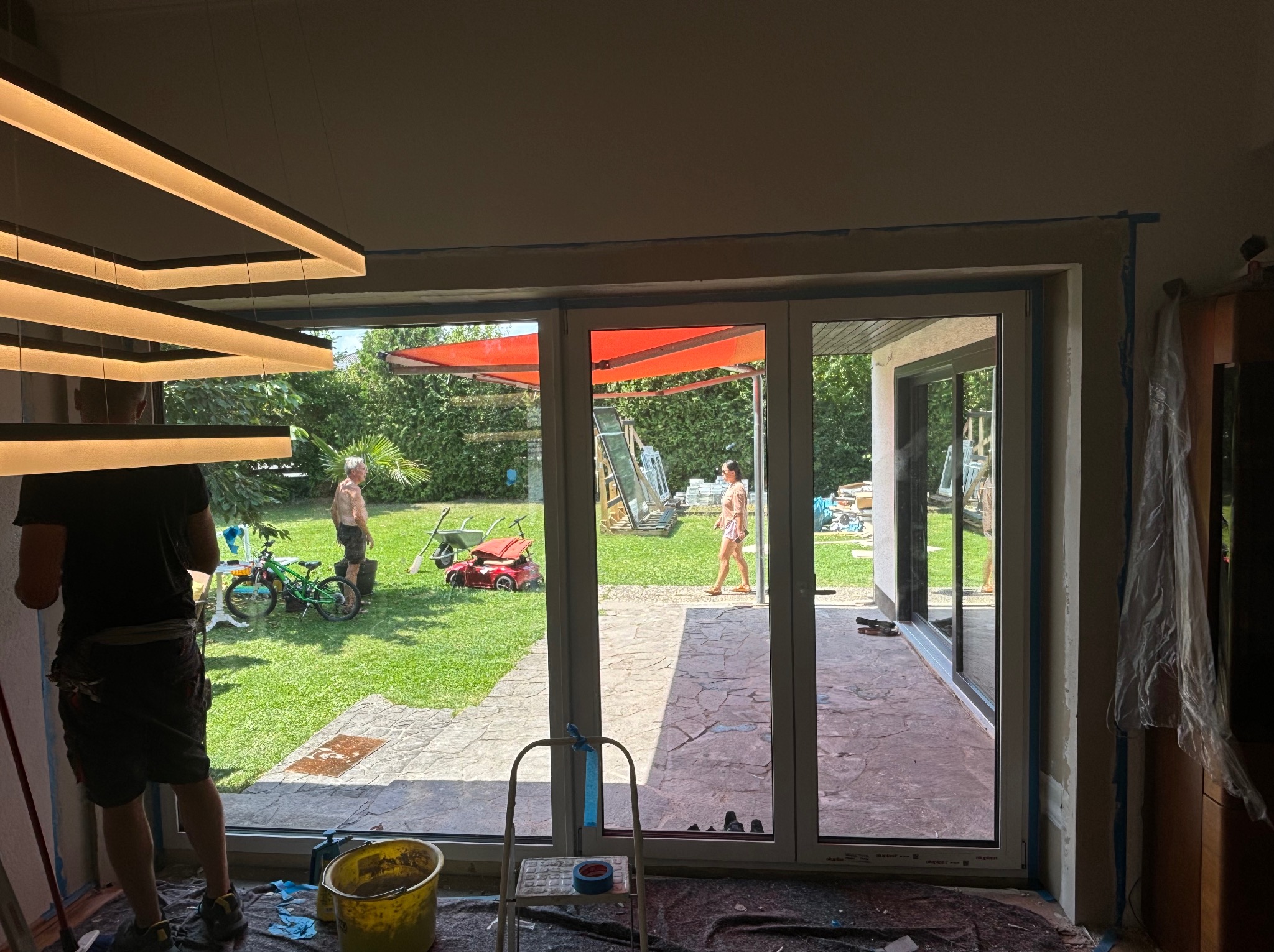 Montaż okna tarasowego: widok na ogród z osobami i sprzętem, widoczne narzędzia i drabina wewnątrz pomieszczenia.