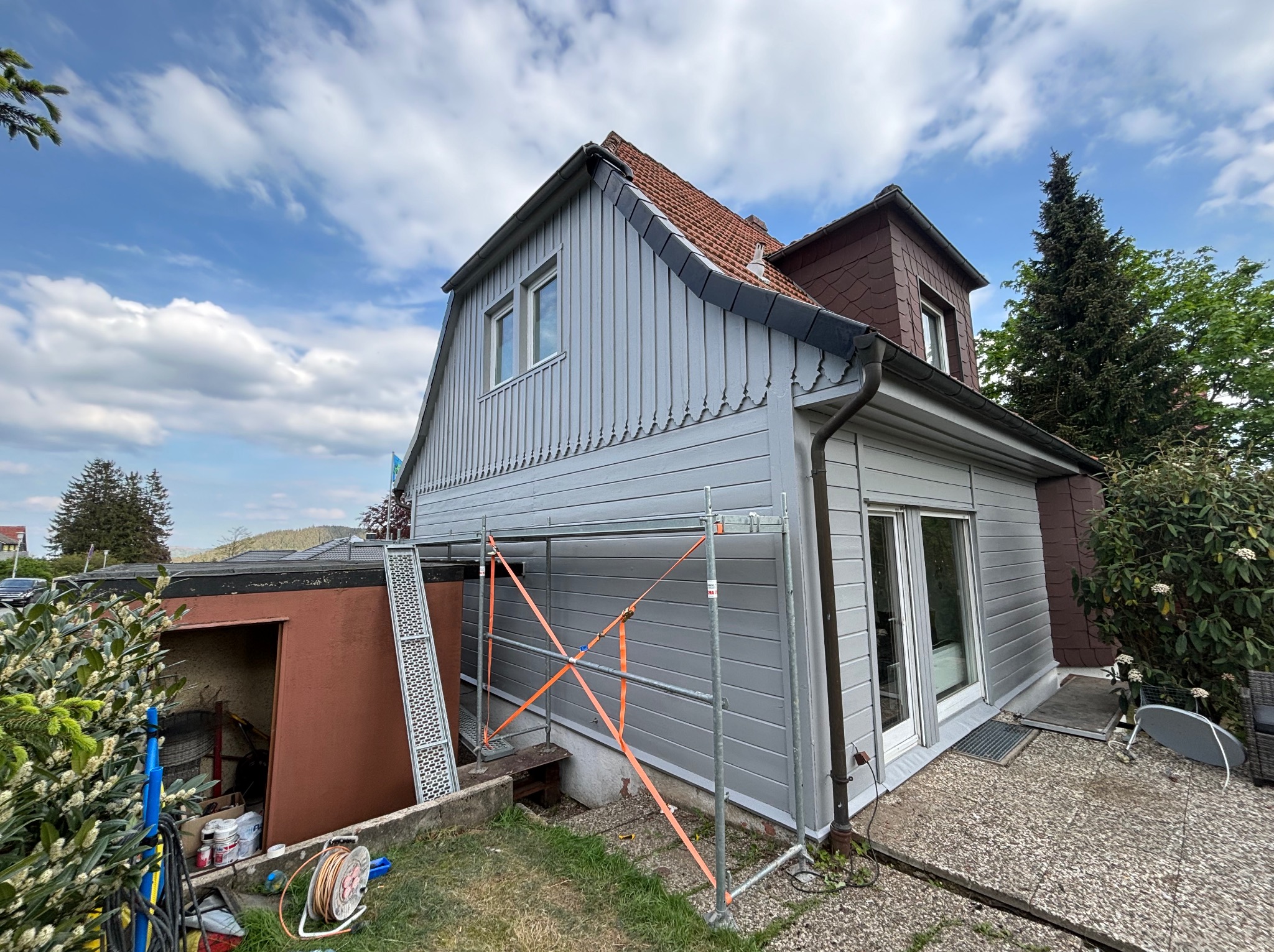 Dom jednorodzinny z szarą elewacją drewnianą, częściowo osłonięty rusztowaniem z pomarańczowymi pasami, widok z niskiej perspektywy na tle błękitnego nieba z chmurami.