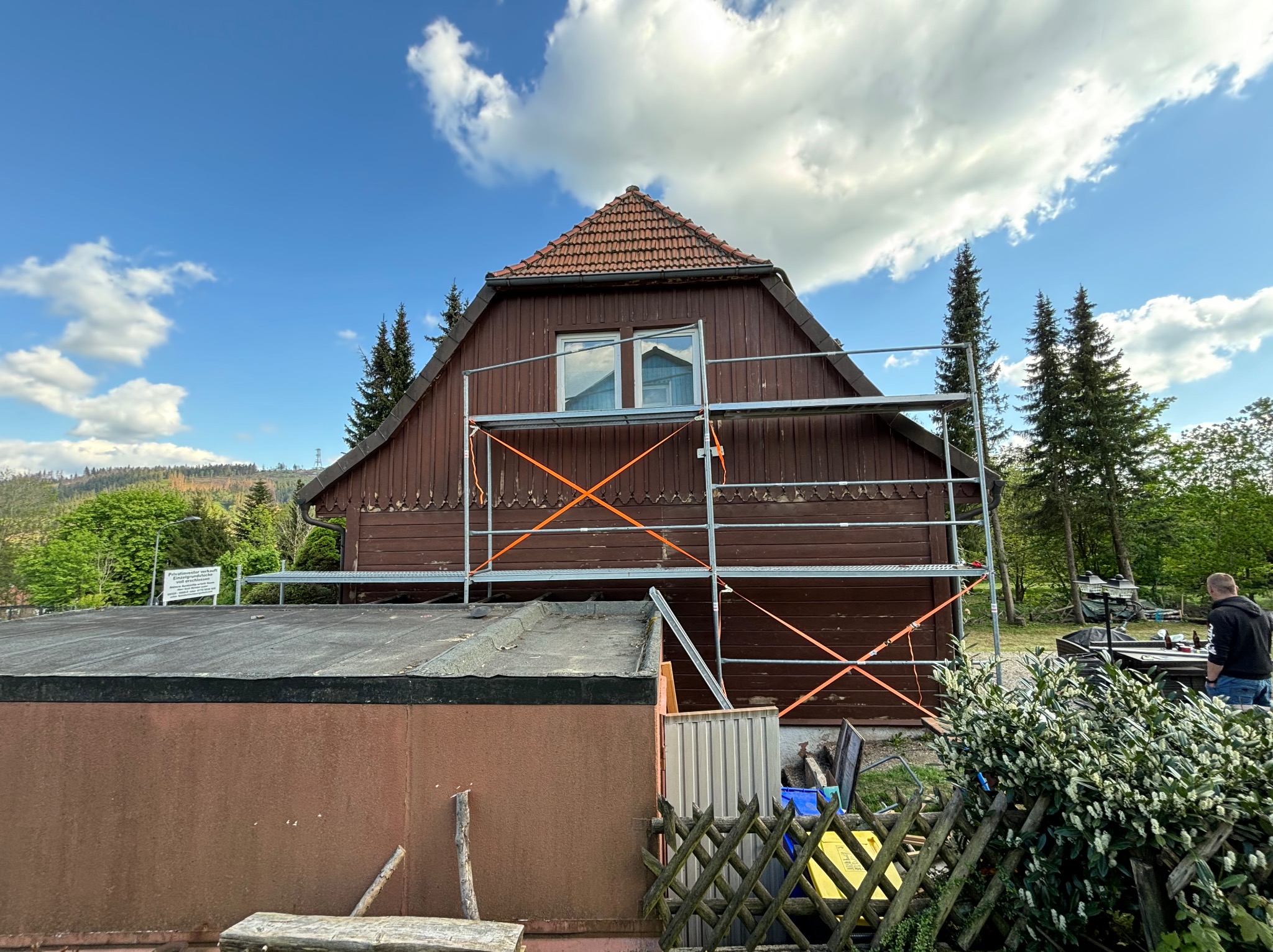 Dwukondygnacyjny dom z drewnianą elewacją, częściowo zakryty rusztowaniem malarskim, z widocznym fragmentem dachu pokrytego czerwoną dachówką i dwoma oknami na poddaszu.