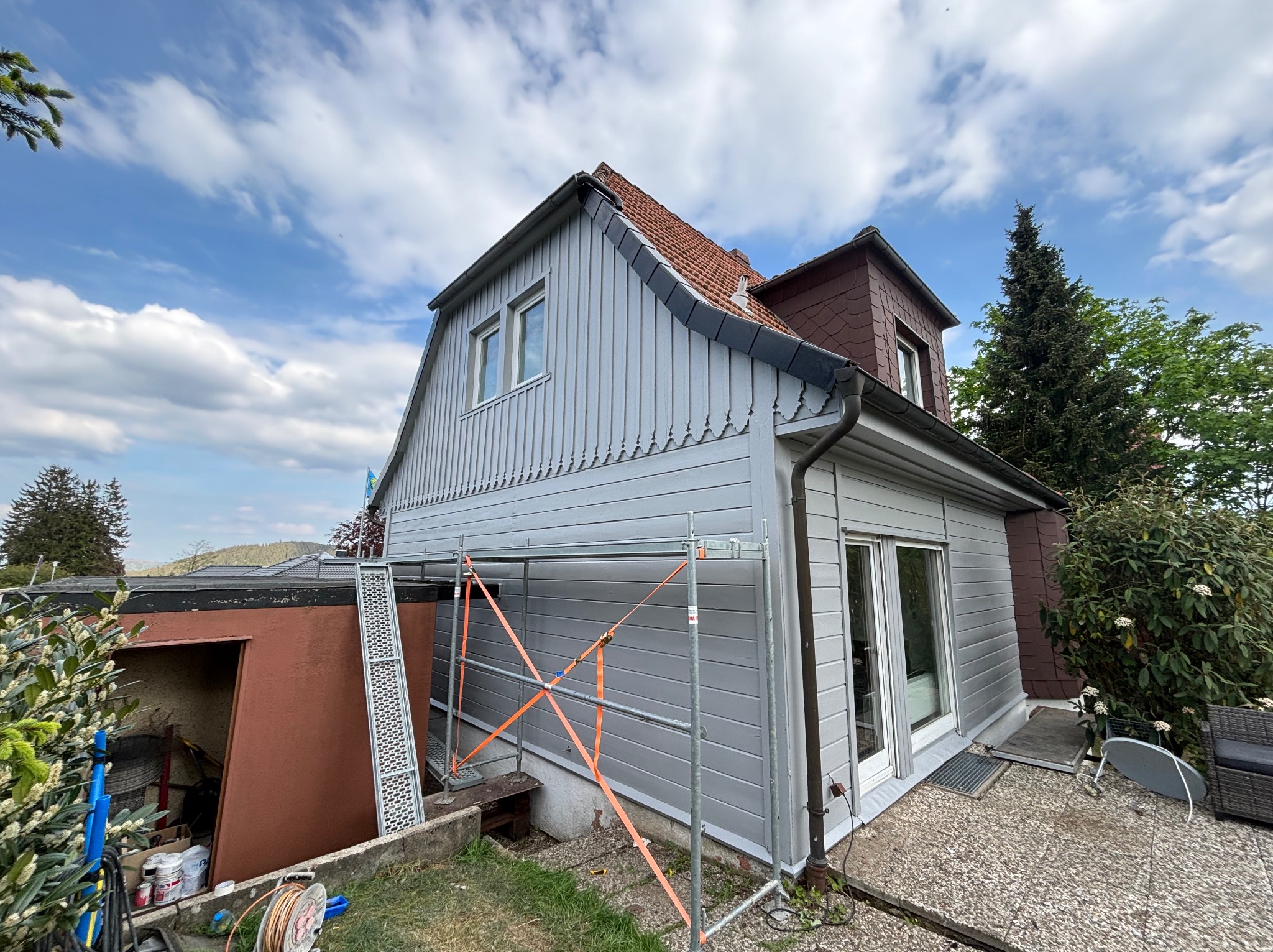 Dom z szarą elewacją w trakcie prac wykończeniowych, widoczne rusztowanie, częściowo odnowiona fasada i dach z czerwonej dachówki.