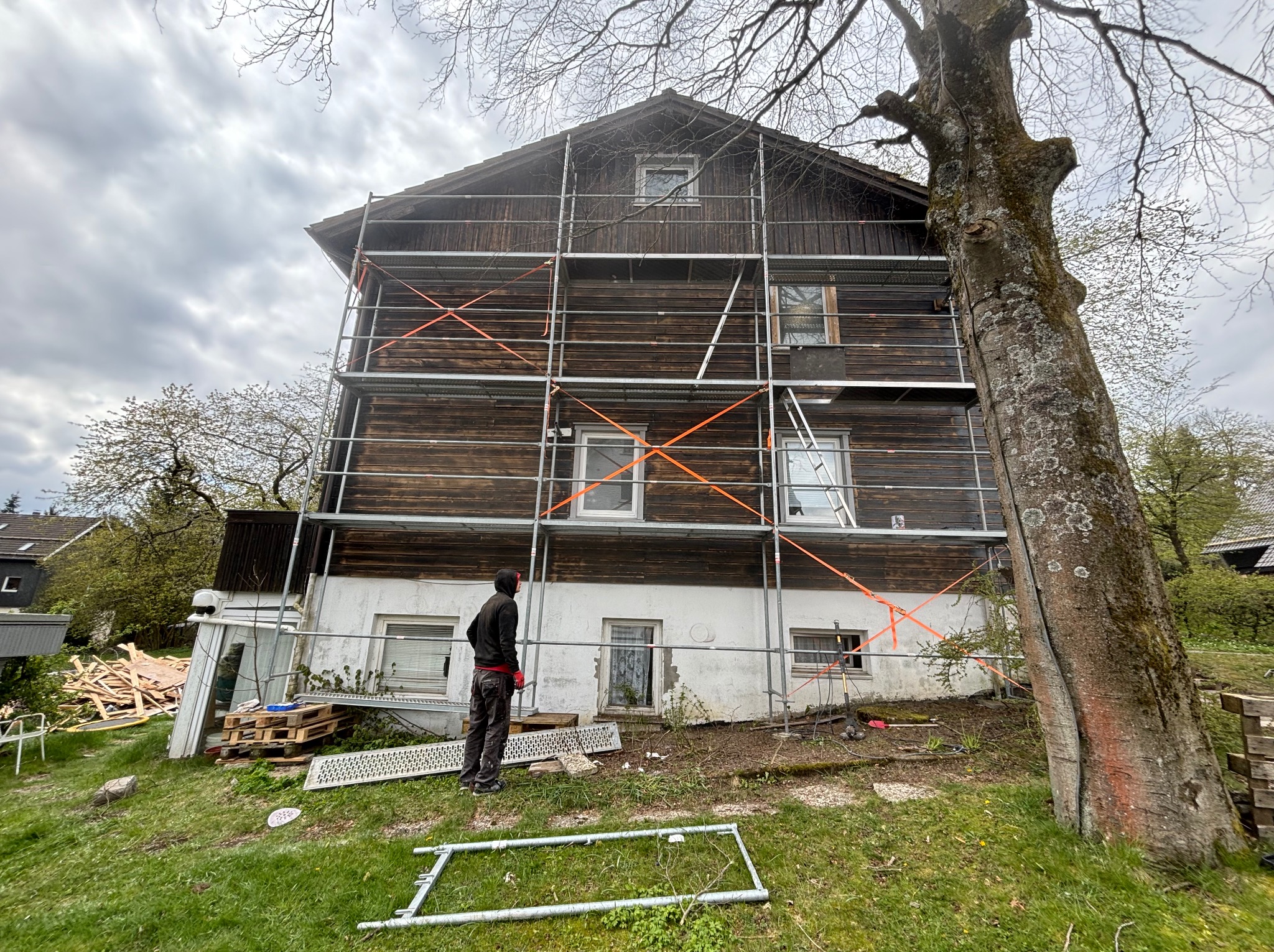 Drewniany dom w trakcie remontu, otoczony rusztowaniem z pomarańczowymi pasami zabezpieczającymi, widoczny pracownik w czarnej bluzie i czerwonych rękawicach, obok leżące deski i duży pień drzewa.