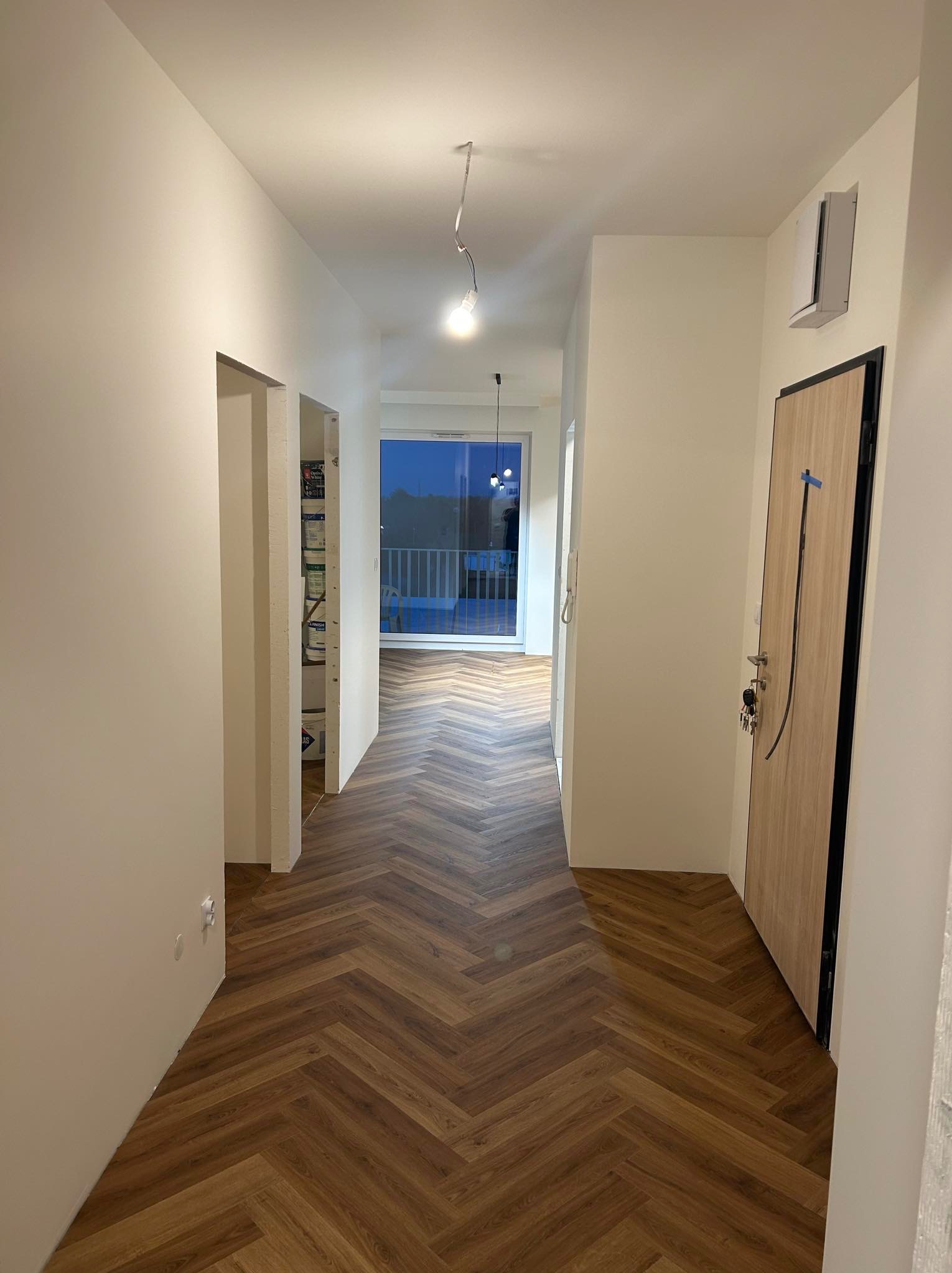 Nowoczesny korytarz z parkietem ułożonym w jodełkę, świeżo pomalowane ściany na biało, widoczne drzwi wejściowe z kluczami i fragment balkonu w tle.