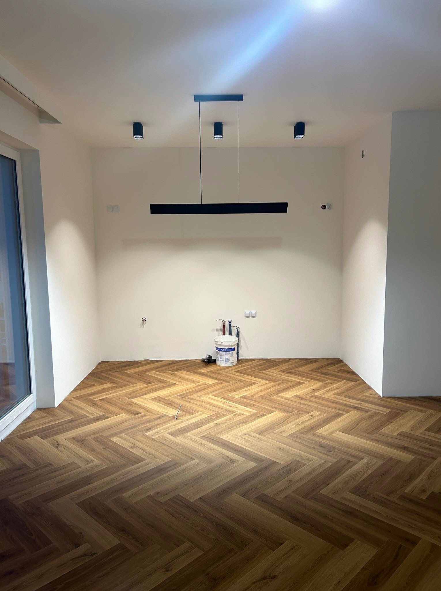 Pusty pokój z ułożonymi panelami w jodełkę, świeżo pomalowanymi ścianami, nowoczesnym oświetleniem sufitowym i widocznym wiadrem z farbą w rogu.