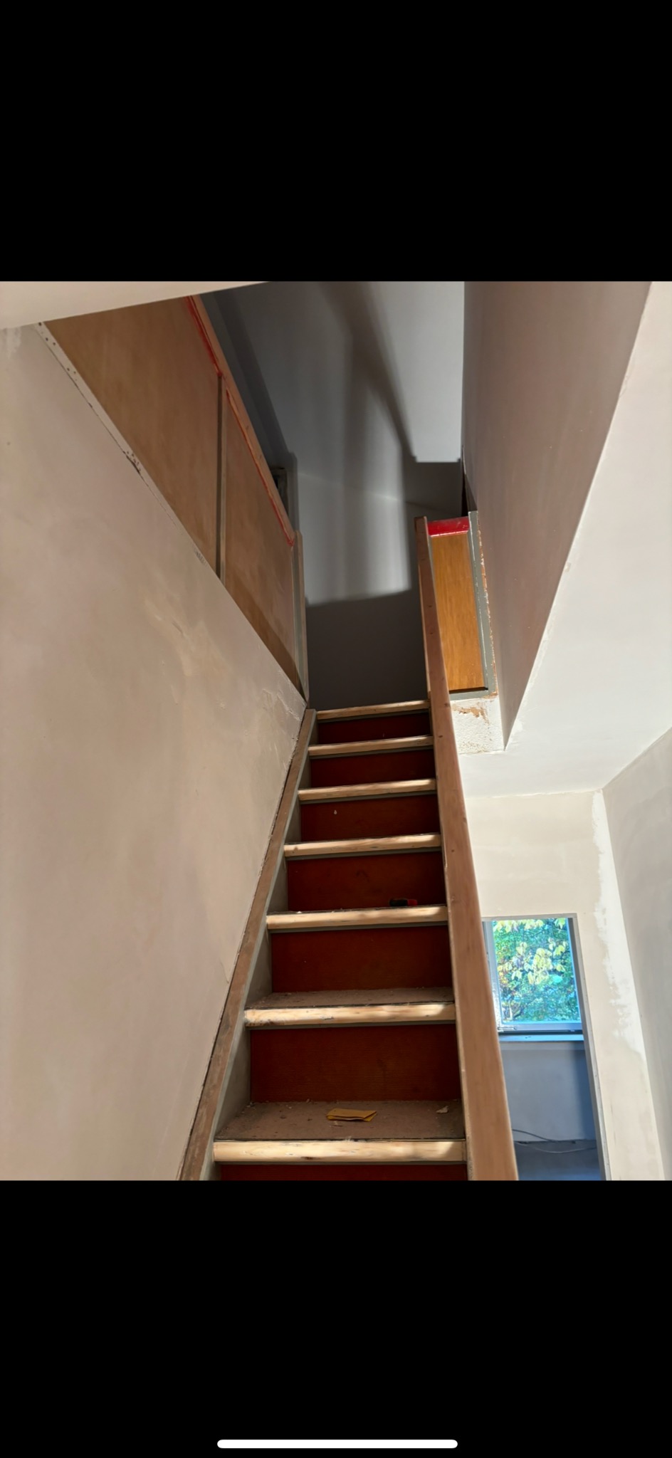 Klatka schodowa w trakcie remontu: ściany po nałożeniu gładzi, drewniane schody z resztkami gruzu, widoczne okno z zielenią na zewnątrz.