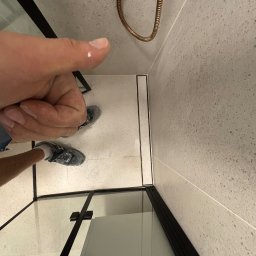 Hrytsiuk - Ukończona kabina prysznicowa z odpływem liniowym, widok z góry, palec wskazujący na kroplę wody na obudowie, szare płytki na ścianach i podłodze.