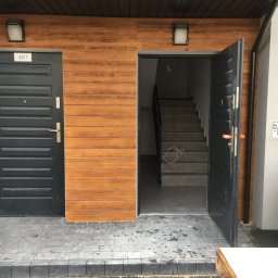 Hrytsiuk - Wejście do budynku z drewnianą elewacją, czarnymi drzwiami z numerem 46/1 i otwartymi drzwiami ukazującymi schody wewnętrzne, widoczne ślady wilgoci na chodniku.