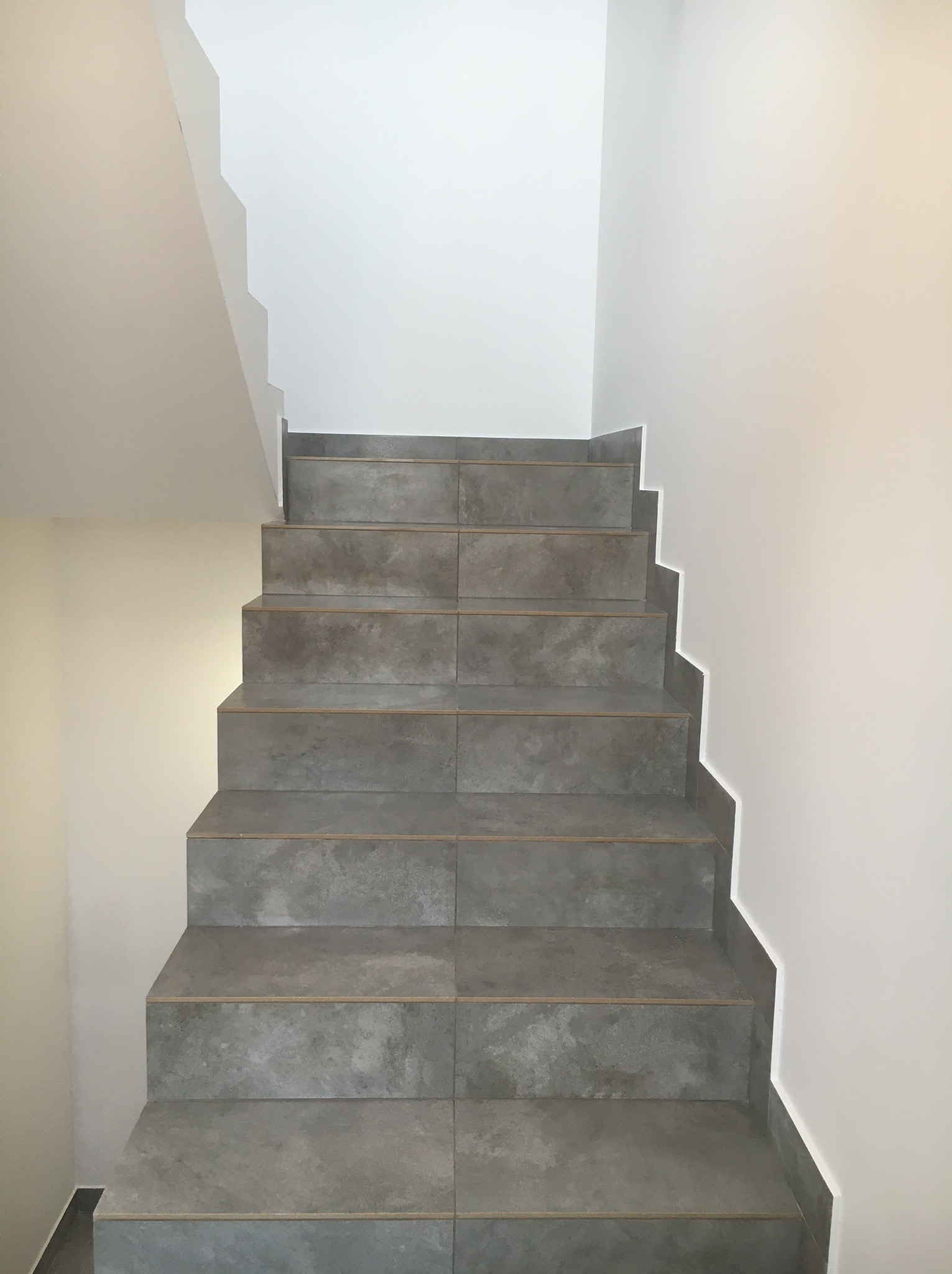 Schody wykończone szarymi płytkami imitującymi beton, prowadzące do góry, z białymi ścianami po bokach. Minimalistyczny design.